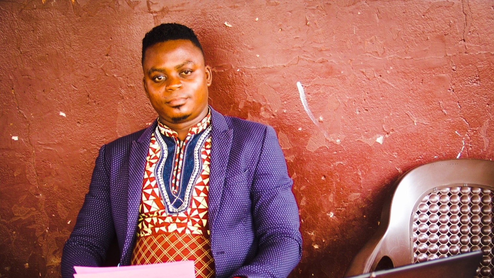 Dans l'image, un homme est assis à une table en bois, près d'un ordinateur portable noir. Il porte une chemise traditionnelle aux motifs colorés, mêlant des nuances de rouge, bleu et blanc, et une veste sombre qui contraste avec sa tenue. Son expression est calme et confiante. Sur la table, on peut voir des feuilles de papier rose ainsi que quelques objets, probablement des lunettes de soleil. Le fond est d'un brun uni, ce qui met en valeur le personnage au premier plan. L'atmosphère semble détendue et conviviale.