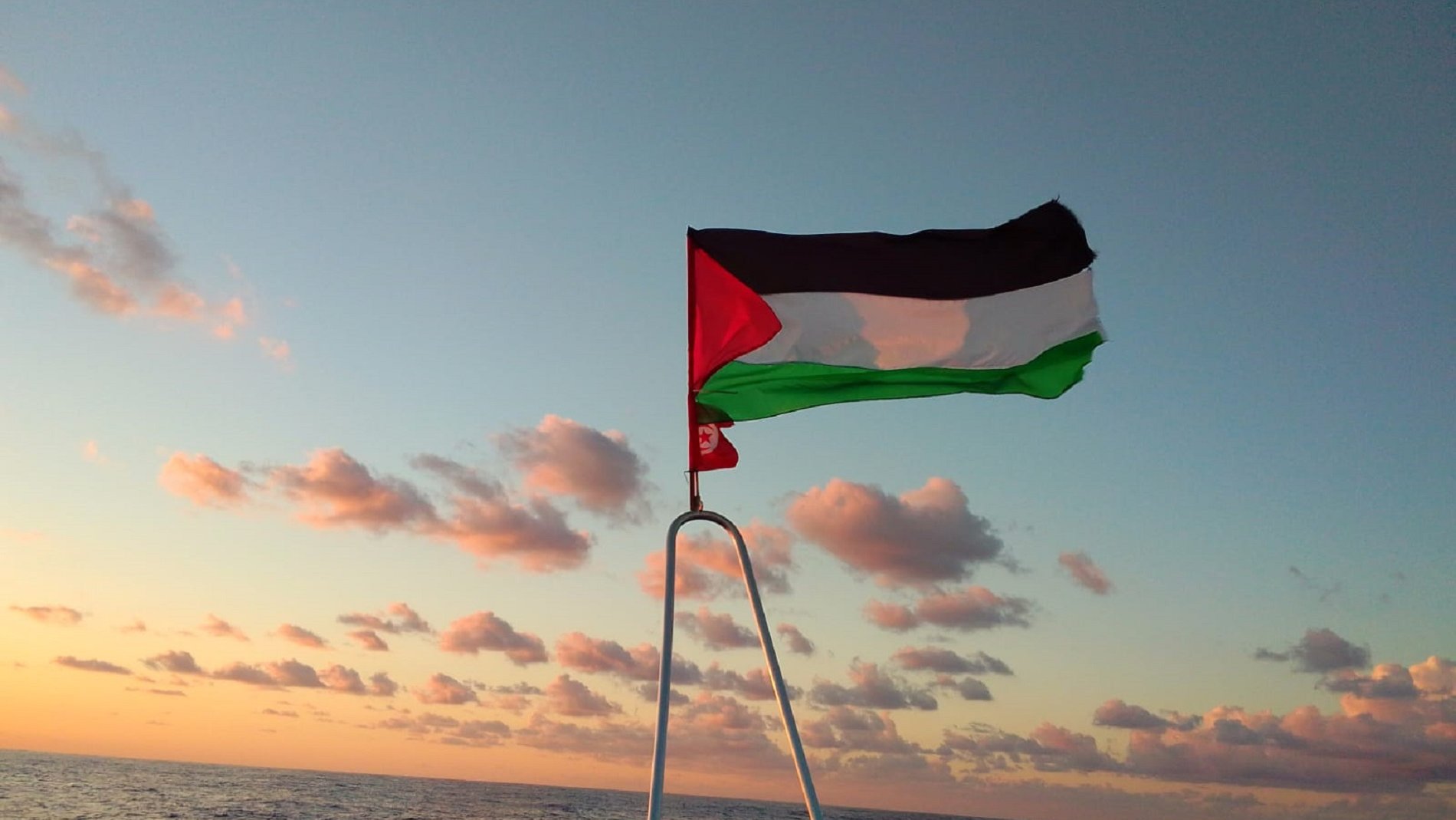 L'image montre un drapeau palestinien flottant sur un mât, avec un vaste horizon marin au fond. Le ciel est teinté de couleurs chaudes, probablement à l'aube ou au crépuscule, avec des nuages épars. Les couleurs du drapeau sont distinctes : le noir, le blanc, le vert et le rouge. La mer est calme et reflète doucement les teintes du ciel, créant une ambiance paisible et solennelle.