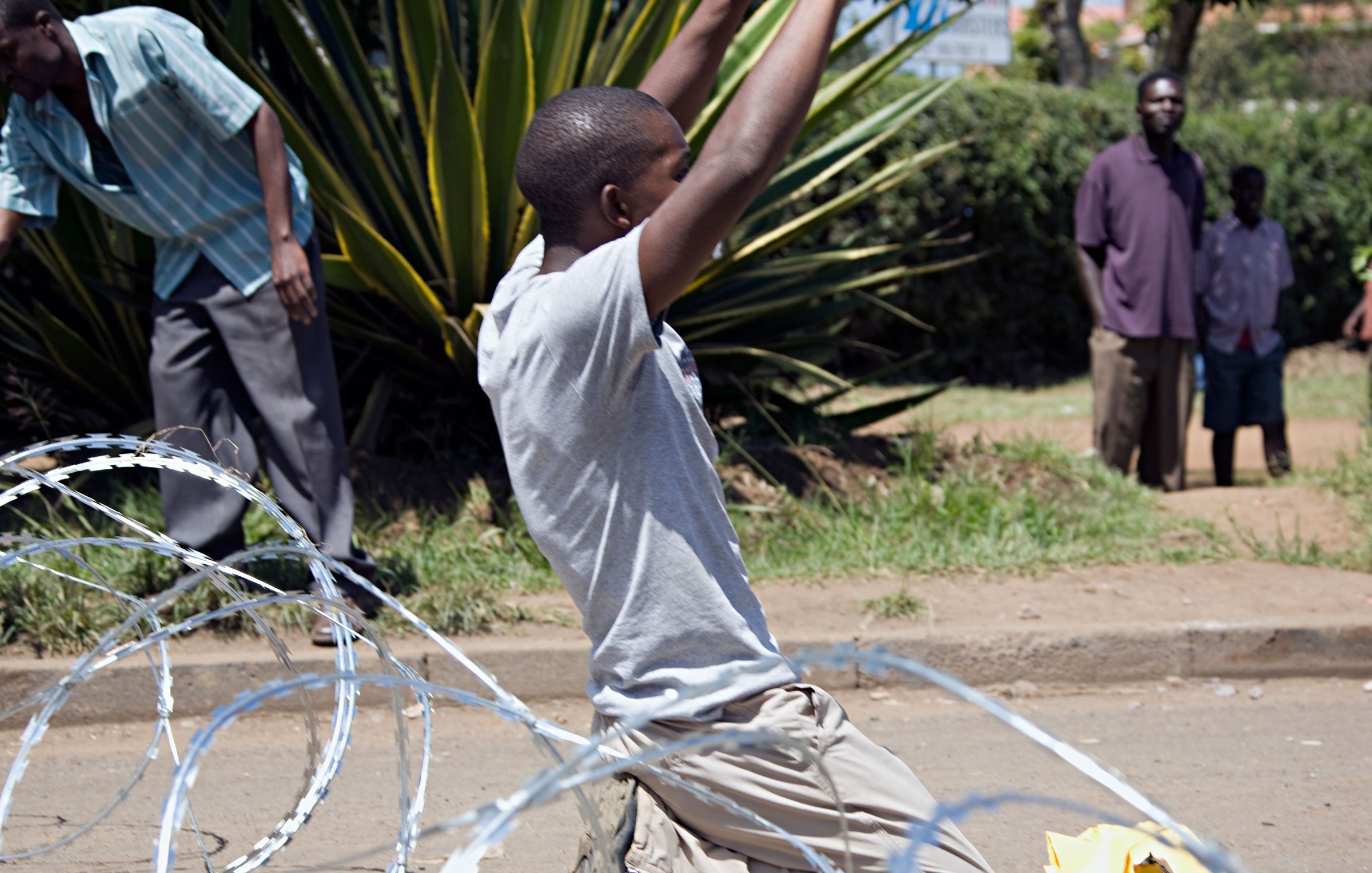 L'image présente une scène animée dans un environnement urbain. Au premier plan, un jeune homme est agenouillé sur le sol, les bras levés dans une posture d'expression intense, peut-être de joie ou de victoire. Il porte un t-shirt gris et un pantalon clair. Autour de lui, des fil de fer barbelés sont dispersés, ajoutant une dimension de tension à la scène. En arrière-plan, plusieurs personnes sont visibles, certaines observant, d'autres engagées dans des activités, suggérant une atmosphère collective. La végétation, avec des plantes vertes, donne un aspect naturel au lieu. L'ensemble de cette image transmet une forte émotion, où les gestes et les postures des individus racontent une histoire d'énergie et de dynamisme.