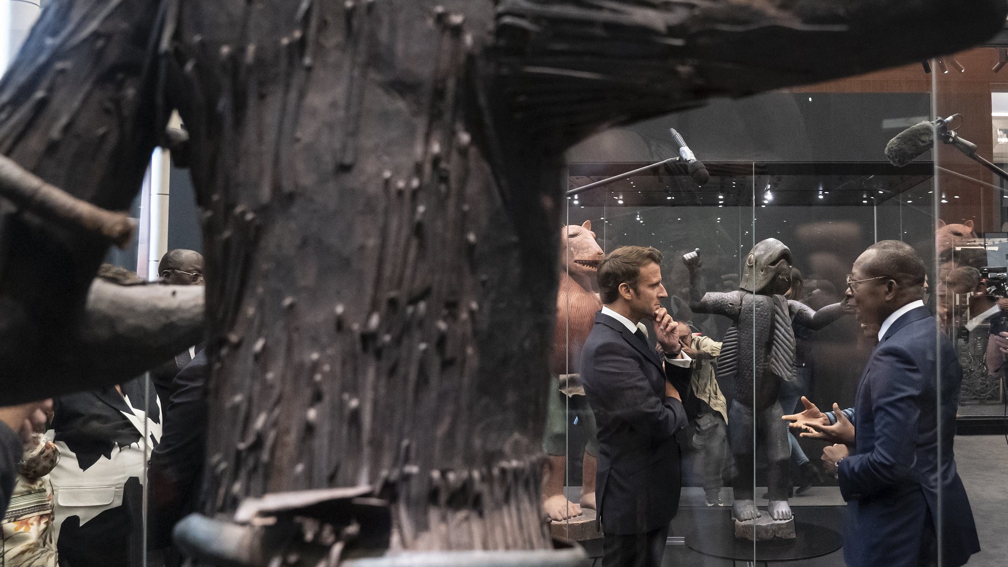 L'image montre un espace d'exposition où deux hommes en costume discutent. L'un d'eux semble concentré sur la conversation, tandis que l'autre écoute attentivement. Ils se tiennent près d'une grande sculpture en bois, qui occupe une place centrale. Cette sculpture a une texture rugueuse, avec des détails qui évoquent une forme humaine ou abstraite. En arrière-plan, d'autres œuvres d'art sont visibles, ajoutant à l'atmosphère culturelle de l'exposition. L'éclairage est doux, mettant en valeur les sculptures tout en créant une ambiance contemplative et respectueuse.