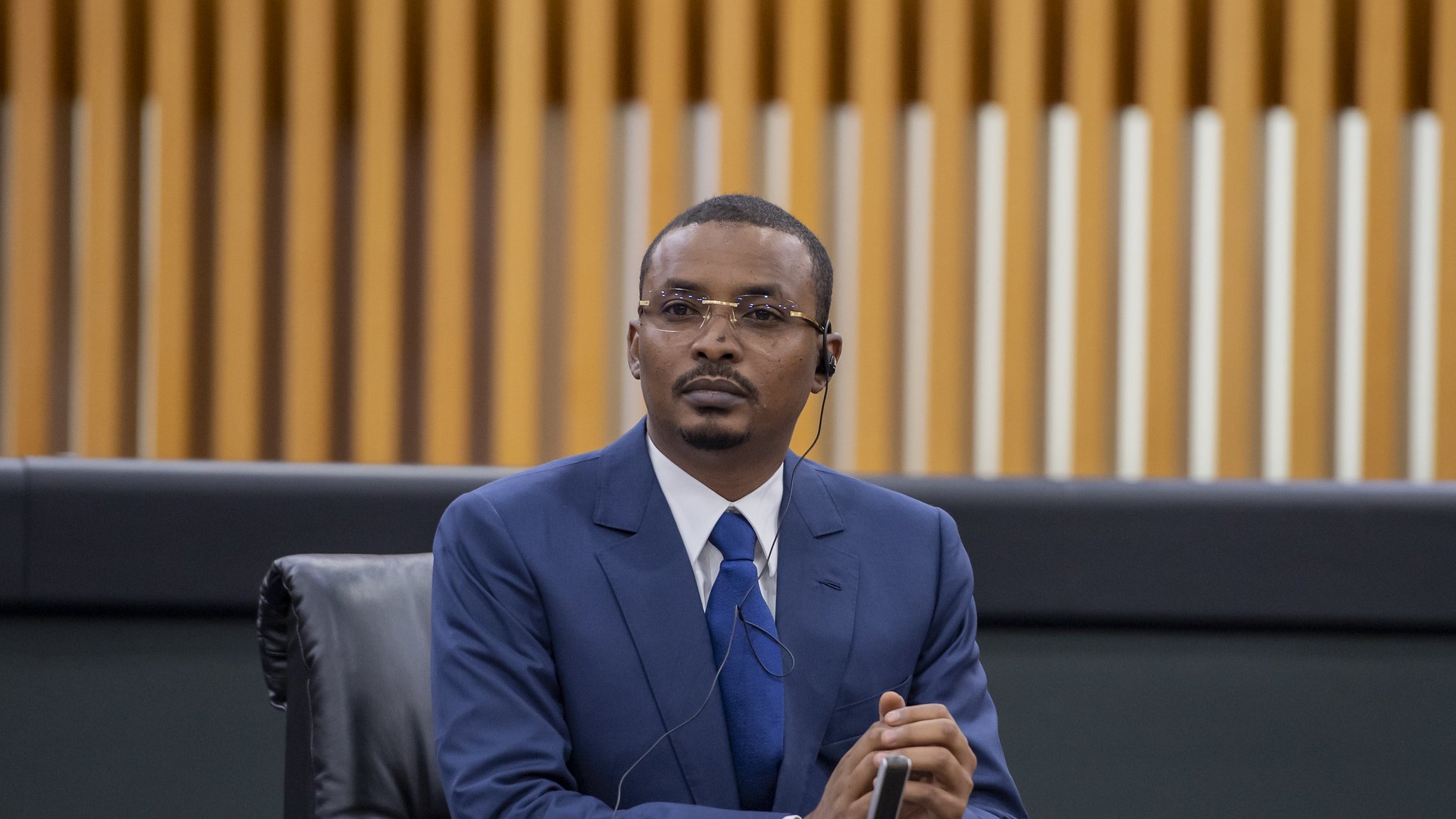 Dans cette image, un homme est assis à une table dans un environnement formel, probablement lors d'une réunion ou d'une conférence. Il porte un costume bleu, une chemise blanche et une cravate bleue. Ses cheveux sont courts et bien coiffés. L'homme semble concentré et attentif, avec un léger froncement de sourcils et les mains croisées devant lui. À son oreille, on peut voir un écouteur, ce qui suggère qu'il écoute probablement une traduction ou une intervention en cours. En arrière-plan, on distingue des panneaux en bois aux lignes verticales, ajoutant une touche moderne à l'espace. L'atmosphère générale est professionnelle et sérieuse.