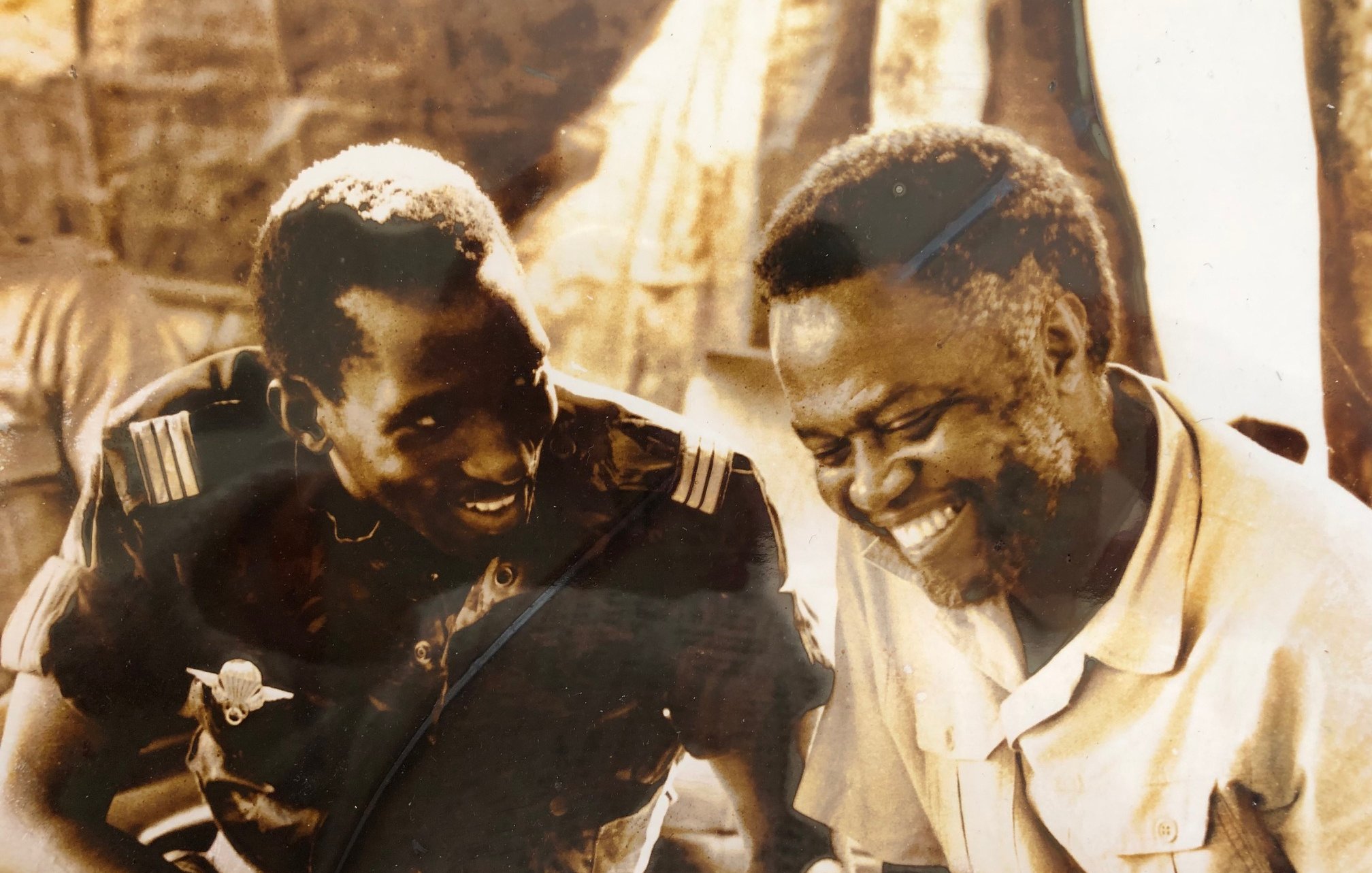 L'image présente deux hommes assis côte à côte, souriant et en train de discuter. L'un d'eux porte un uniforme militaire, tandis que l'autre est habillé d'une chemise claire. Leurs visages expriment la joie et la camaraderie. En arrière-plan, on peut distinguer des silhouettes floues de personnes et de la végétation, ce qui suggère un environnement social dynamique. Les teintes de l'image sont sépia, ce qui lui donne un aspect vintage, évoquant une époque passée.
