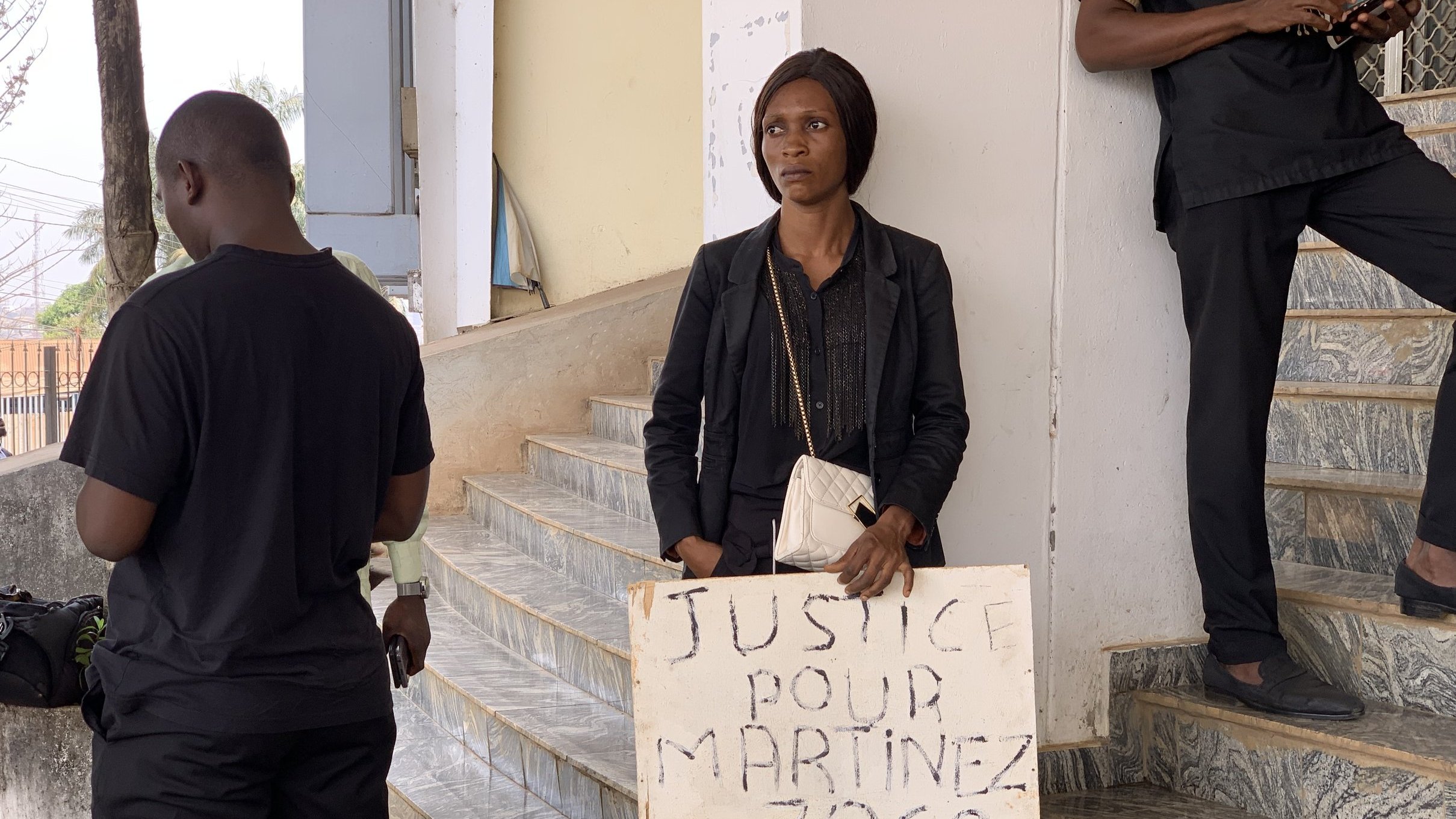 Dans cette image, on voit une femme debout dans un espace public. Elle porte des vêtements noirs et tient un panneau en bois qui porte l'inscription "JUSTICE POUR MARTINEZ ZOGO". La femme a une expression sérieuse et semble déterminée. À proximité, plusieurs personnes en vêtements noirs se tiennent, suggérant un rassemblement ou une manifestation. L'environnement est urbain, avec des escaliers visibles et des bâtiments en arrière-plan, indiquant un cadre où des actions sociales ou des revendications ont lieu.