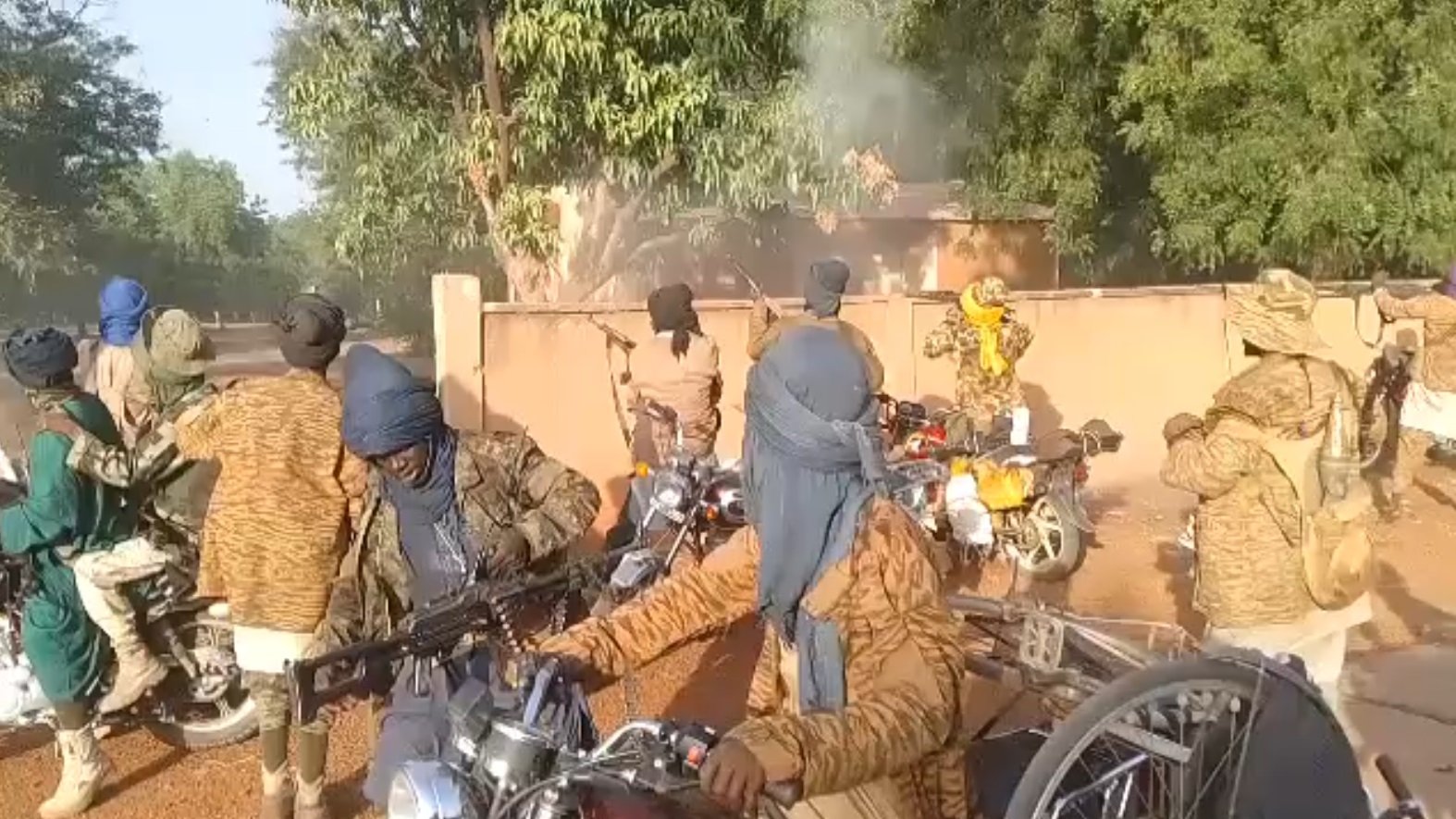 L'image montre un groupe de personnes sur des motos, portant des vêtements qui masquent leur visage et leur tête, probablement pour des raisons de camouflage ou de protection. Ils semblent être dans un environnement extérieur, avec des arbres et un bâtiment en arrière-plan. Il y a de la fumée qui s'échappe, indiquant peut-être une situation de tension ou d'activité intense. Certains individus tiennent des armes, ajoutant une atmosphère de gravité à la scène. Les motos sont variées et leur position suggère qu'ils sont en mouvement ou prêts à partir.