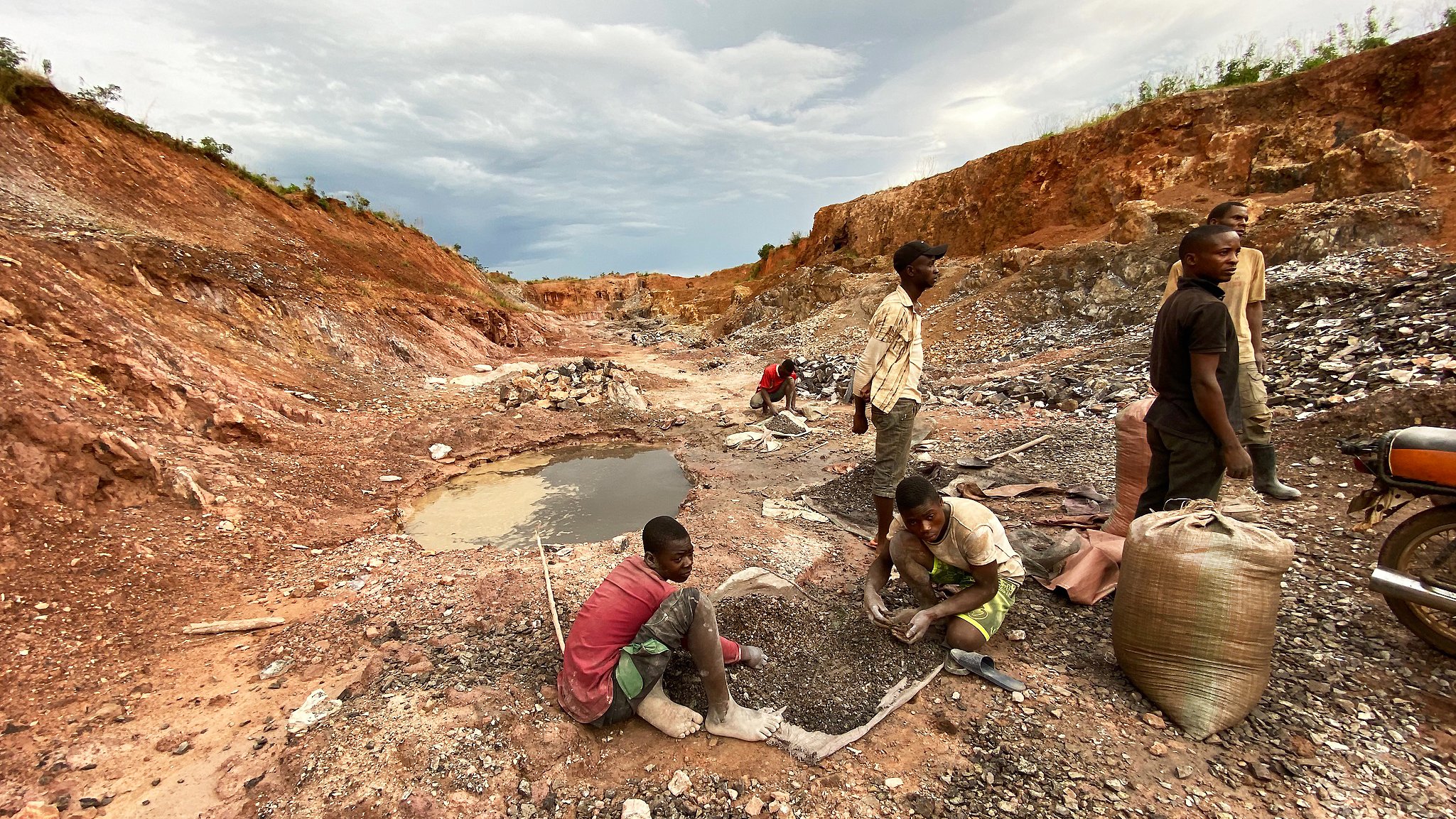 L'image montre une scène rurale d'exploitation minière. On y voit un paysage marqué par des fouilles, avec un sol rouillé et des montagnes de terre séparées. Au premier plan, plusieurs personnes, principalement des hommes et des jeunes garçons, sont en train de travailler. Certains sont accroupis, tamisant des pierres ou ramassant des minéraux, tandis que d'autres semblent transporter des sacs pleins de matériaux. À gauche, il y a un petit plan d'eau, qui est trouble, entouré de débris miniers. Le ciel est nuageux, ajoutant une ambiance sombre à la scène, évoquant un travail difficile et épuisant.