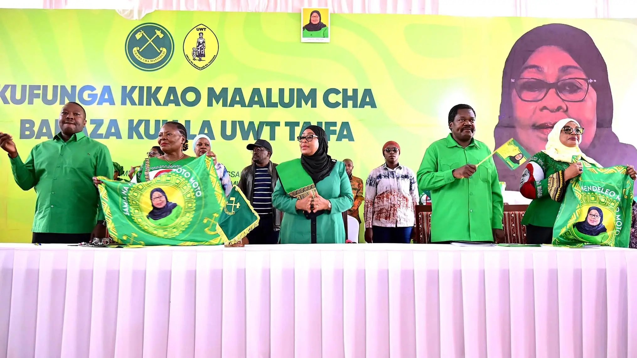 L'image montre un événement politique avec plusieurs personnes debout derrière une table, vêtues de chemises vertes. Au fond, on peut voir une grande bannière avec un portrait d'une femme souriante, probablement une dirigeante. Les participants tiennent des drapeaux ou des banderoles, souvent en vert et jaune, qui portent des symboles liés à leur organisation. L'atmosphère semble festive et engagée, avec des membres du public en arrière-plan qui écoutent attentivement. La réunion semble évoquer des thèmes de solidarité et de mobilisation.