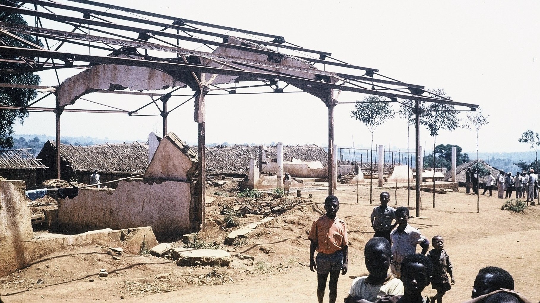 L'image montre un paysage de dévastation avec des structures en ruine. À l'avant-plan, plusieurs personnes, dont des enfants et des adultes, se tiennent debout sur un sol poussiéreux. En arrière-plan, on voit des bâtiments partiellement effondrés, avec des toits manquants et des murs endommagés. Des arbres isolés se dressent ici et là, ajoutant une touche de verdure à ce cadre désolé. Le ciel est dégagé et ensoleillé, contrastant avec l'état des lieux. L'atmosphère suggère une scène de désolation, peut-être après un événement tragique.