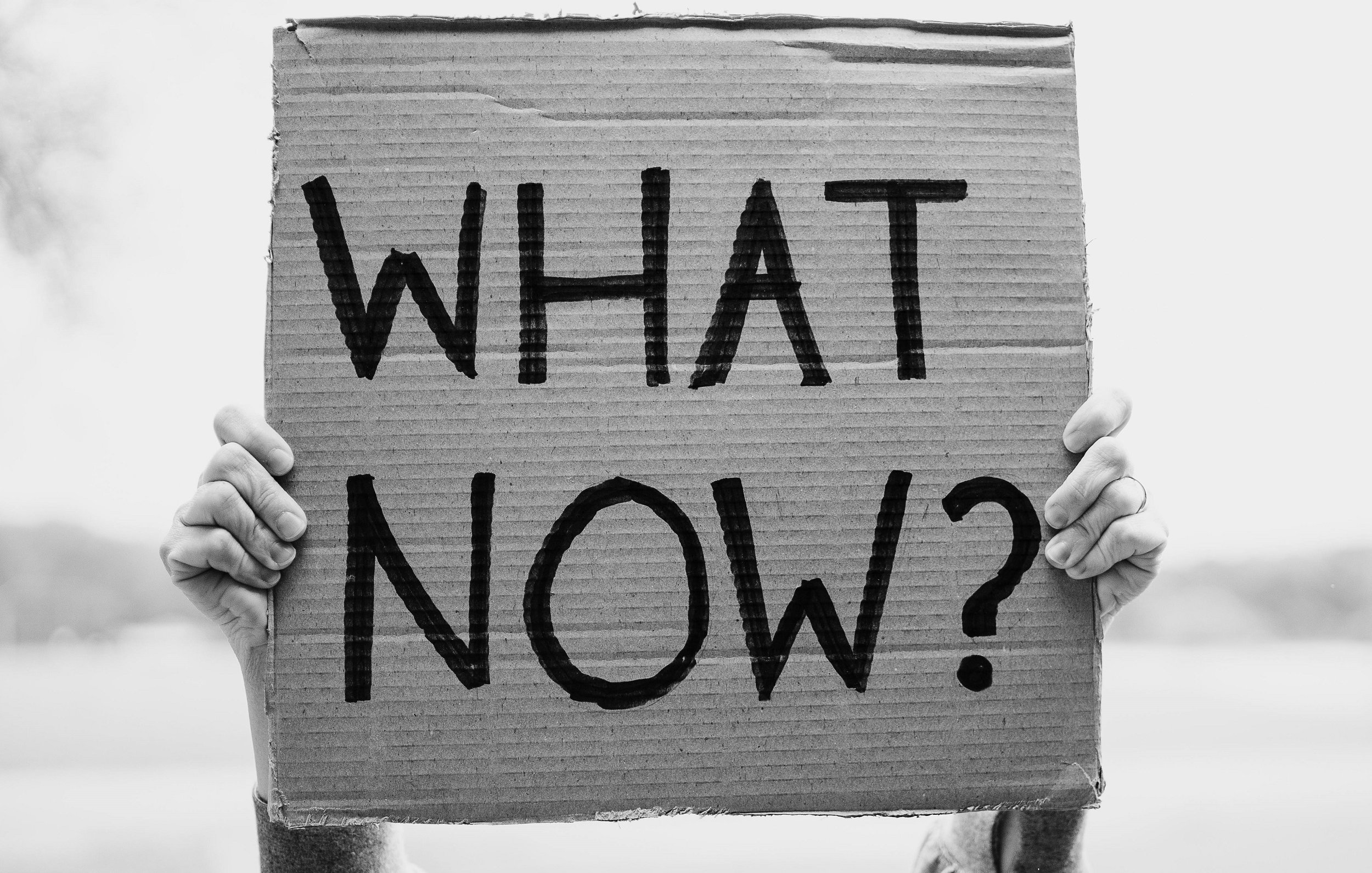 L'image montre une personne tenant une grande pancarte en carton. La pancarte est de couleur marron et comporte des lettres noires écrites en gras. Les mots affichés sont "WHAT NOW ?", ce qui peut exprimer une question ou une incertitude. L'arrière-plan semble flou, donnant un effet de profondeur et mettant en avant le message de la pancarte. La scène est en noir et blanc, ce qui accentue la simplicité et l'impact du message.
