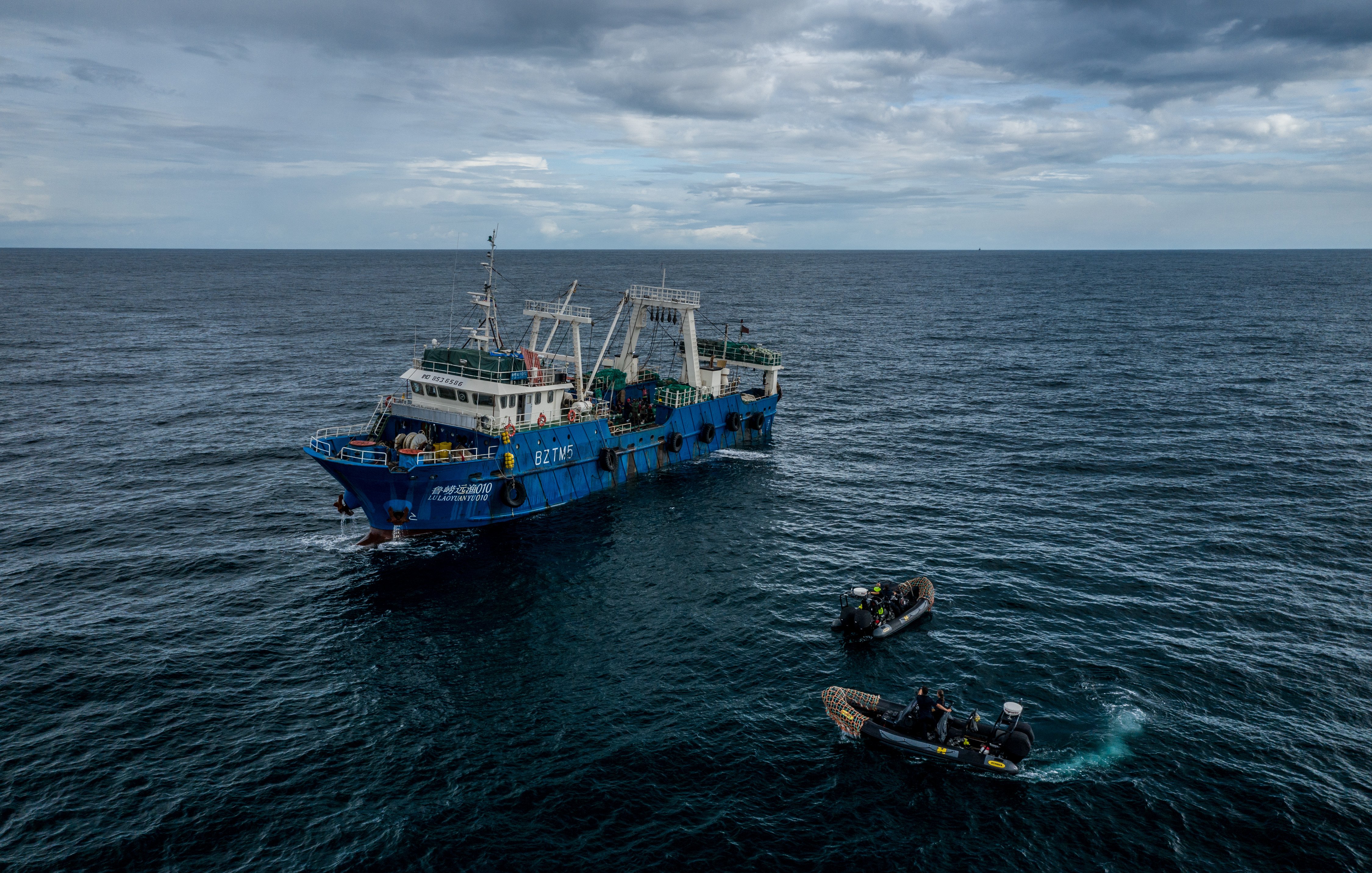 L'image montre un grand navire de pêche de couleur bleue, flottant au milieu de l'océan. Le bateau est en mouvement sur des eaux agitées, sous un ciel nuageux qui laisse présager une légère brise. À côté du navire principal, on peut voir deux petites embarcations, probablement des canots, qui sont en train de s'approcher. Les membres d'équipage, habillés en tenues de plongée, semblent occupés à des activités liées à la pêche. L'ambiance générale dégage une impression de travail maritime, avec le son des vagues et un léger souffle du vent.