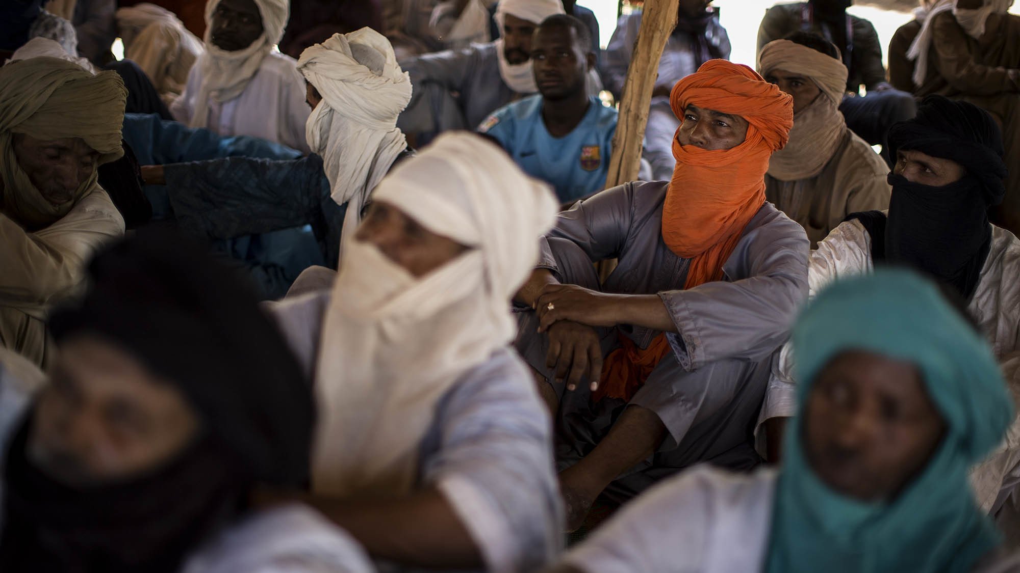 L'image montre un groupe de personnes assises dans un espace clos. Les individus portent des vêtements traditionnels, souvent en tissu léger, et sont partiellement masqués par des turbans ou d'autres pièces de tissu. On peut percevoir des couleurs variées : certains portent des turbans orange, d'autres bleus ou verts. Les visages des personnes sont en grande partie cachés, créant une atmosphère de mystère. L'environnement semble calme, et les positions des corps indiquent une attention collective. La lumière est douce, suggérant un éclairage naturel venant d'une source extérieure. On peut également sentir une ambiance de rassemblement, possiblement pour un échange culturel ou une discussion importante.