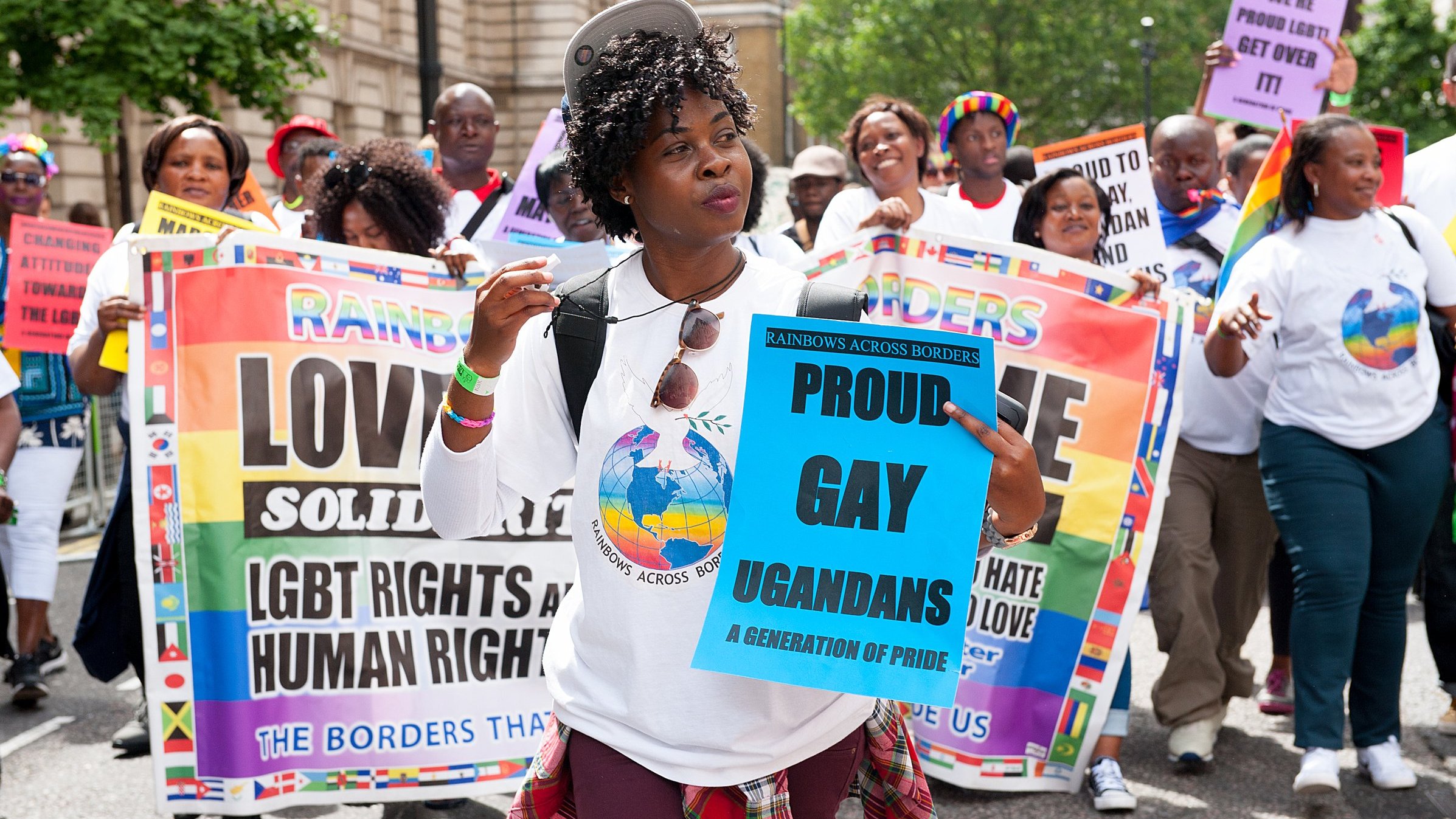 Sur cette image, un groupe de personnes marche ensemble lors d'une manifestation. Au premier plan, une femme souriante porte un t-shirt blanc avec un logo coloré représentant un globe. Elle tient un panneau bleu sur lequel il est écrit "PROUD GAY UGANDANS" en lettres grandes et claires. En arrière-plan, d'autres participants portent divers panneaux affichant des messages de solidarité et de revendication des droits LGBTQ+. La scène est animée et colorée, avec des drapeaux et des tenues qui soulignent la diversité et la fierté. L'ambiance est festive, mais également engagée, reflétant une atmosphère de lutte pour les droits humains.