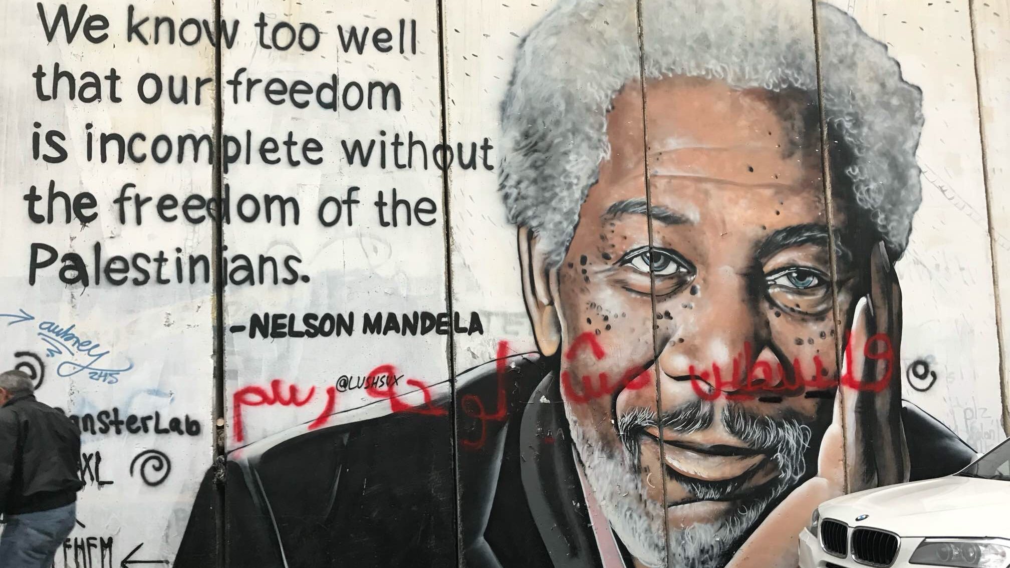 L'image représente un mur sur lequel est peint un mural poignant. À gauche, on voit un portrait détaillé d'un homme âgé avec des cheveux grisonnants et une expression réfléchie. À droite du portrait, une citation attribuée à Nelson Mandela évoque l'importance de la liberté des Palestiniens pour garantir notre propre liberté. Le texte est en anglais, avec un style graphique qui attire l'attention. En bas, quelques graffitis ajoutent une touche de couleur et de dynamisme à l'ensemble. L'ambiance est à la fois engageante et réfléchie, signifiant une connexion profonde entre les luttes pour la liberté.