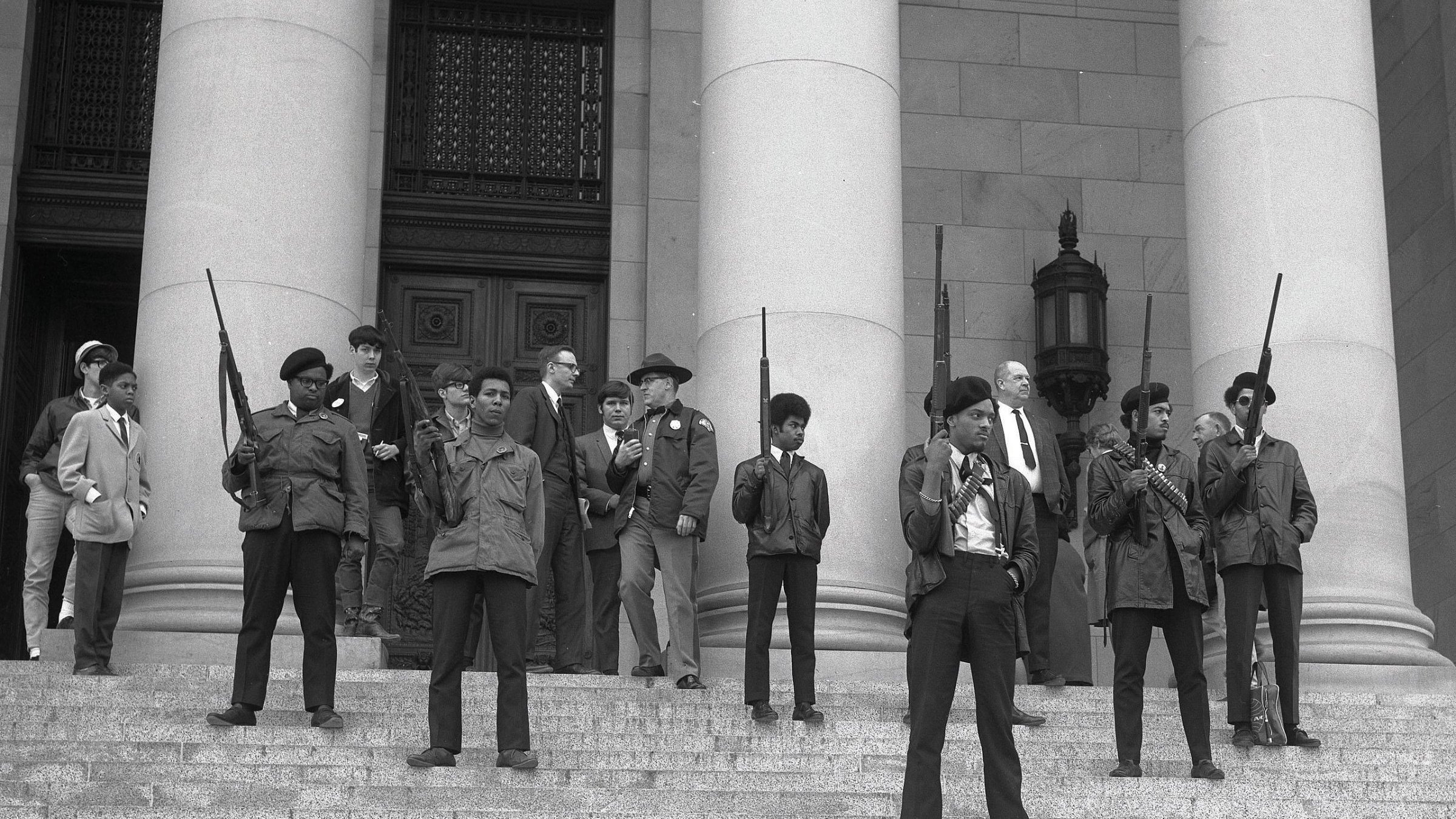 L'image montre un groupe de personnes sur les marches d'un bâtiment imposant, probablement une institution gouvernementale. Au premier plan, plusieurs jeunes hommes se tiennent debout, portant des vestes sombres et des armes à feu, avec des expressions déterminées sur leurs visages. Ils sont entourés d'autres individus, certains vêtus de manière formelle, comme des costumes et des chapeaux. Tous semblent engagés dans une sorte de manifestation ou de rassemblement, émettant un sentiment de tension et de détermination. L'architecture du bâtiment est majestueuse, avec de grandes colonnes en arrière-plan. L'atmosphère générale de l'image évoque une période de lutte pour les droits civiques.