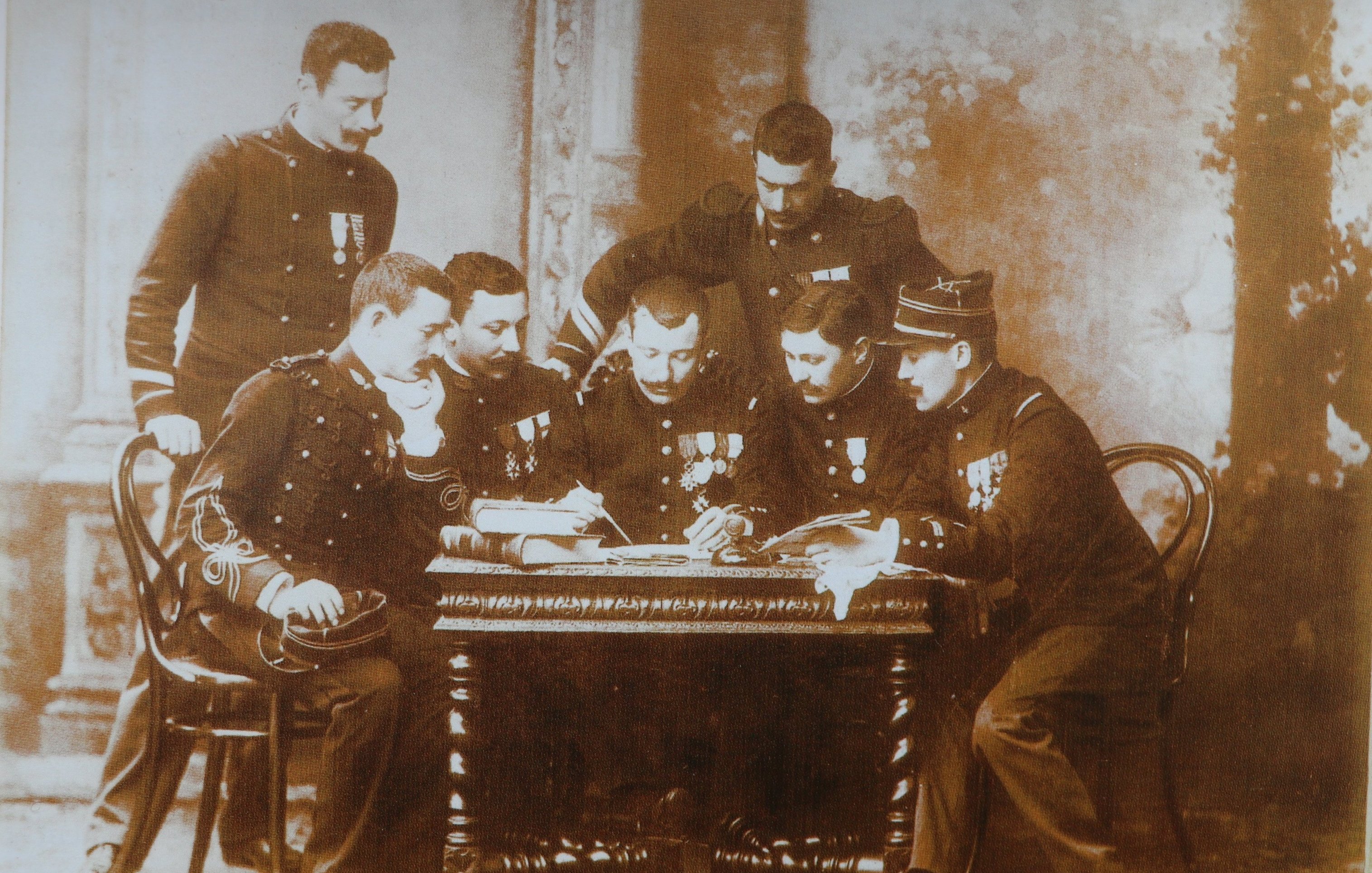 Cette image en noir et blanc représente un groupe de six hommes en uniforme, assis autour d'une table en bois. Ils semblent être des membres d'un corps militaire, chacun portant des médailles décoratives sur leur uniforme. Les hommes sont concentrés sur un document ou un plan posé sur la table, et leur posture indique un échange d'idées ou de discussions sérieuses. En arrière-plan, on peut distinguer quelques éléments architecturaux flous, suggérant un intérieur de bâtiment classique. L'atmosphère se dégage une certaine solennité et camaraderie.