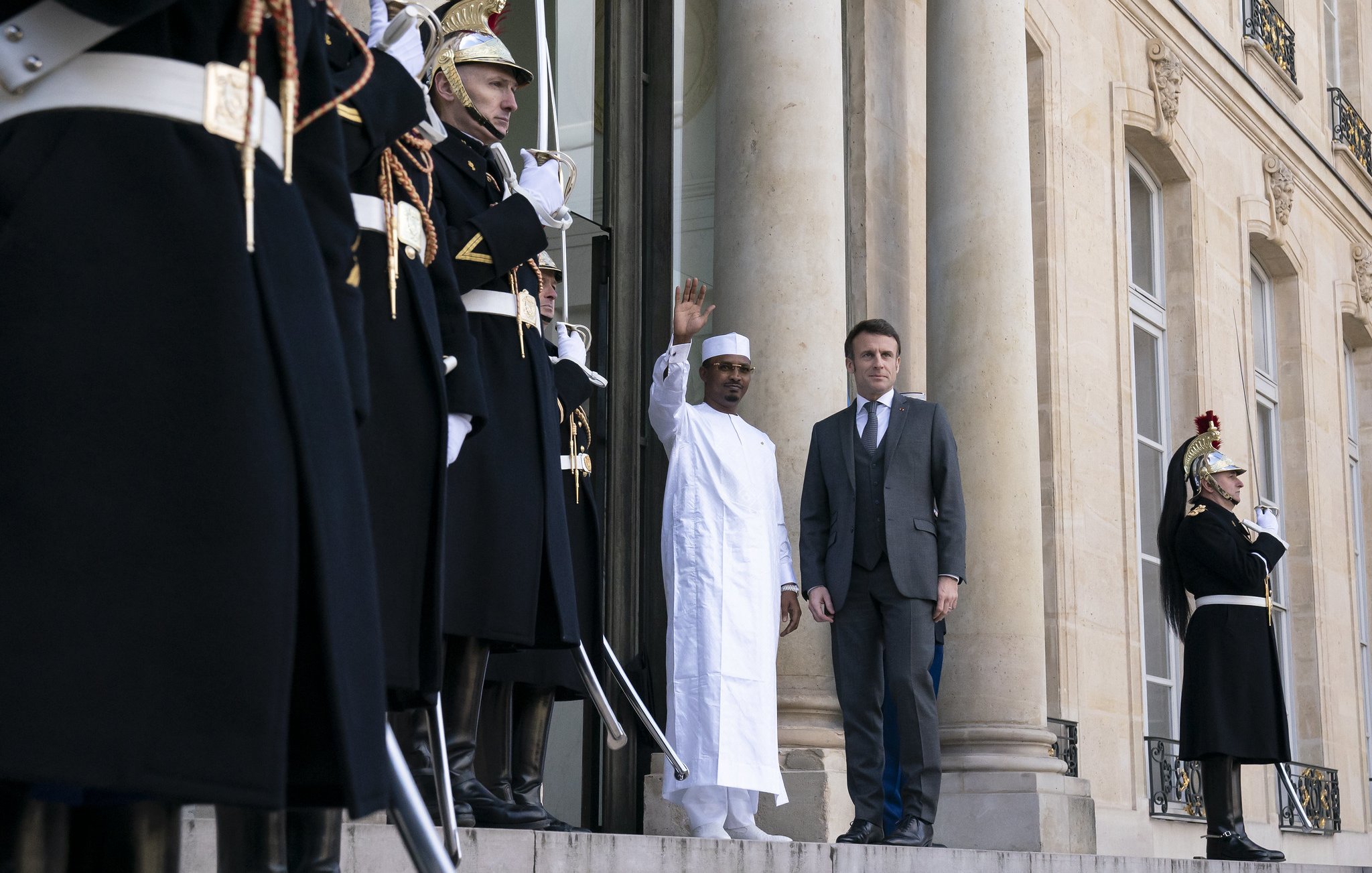 L'image montre une scène officielle à l'extérieur d'un bâtiment majestueux. Deux hommes se tiennent sur les marches devant l'entrée principale. À gauche, un homme porte une tenue traditionnelle blanche, levant la main en salutation. À sa droite, un homme en costume sombre regarde légèrement vers lui. En arrière-plan, plusieurs gardes d'honneur, habillés de costumes noirs avec des ornements dorés et des plumes rouges sur leurs casques, se tiennent en position, ajoutant à l'atmosphère solennelle de la scène. Les colonnes imposantes du bâtiment et les détails architecturaux mettent en valeur le caractère formel de l'événement.