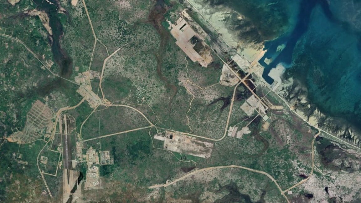 L'image représente une vue aérienne d'une région dont le paysage est marqué par des routes, des structures et des zones déforestées. On peut observer des chemins sinueux qui relient différents points, ainsi que des zones construites qui semblent être des installations industrielles ou militaires. La présence de la mer à droite de l'image ajoute un contraste avec la verdure terrestre, et on peut également voir des étendues d'eau et des zones côtières. Les nuances de couleur vont du vert des zones boisées aux teintes plus claires des infrastructures.