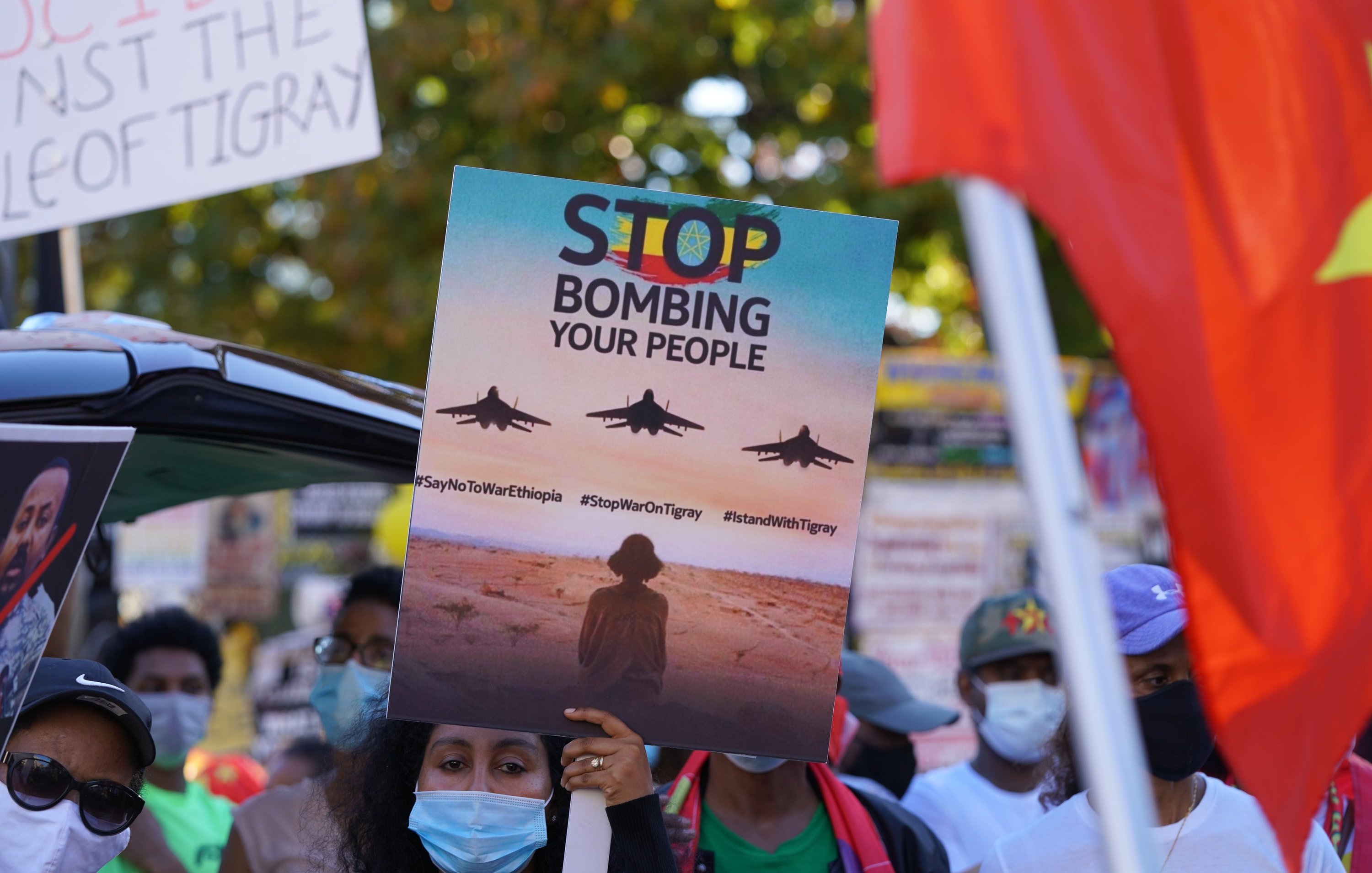 L'image montre une scène de manifestation. Au premier plan, une personne tient une grande pancarte qui affiche le message "STOP BOMBING YOUR PEOPLE" avec des images d'avions de chasse en vol. La personne est entourée d'autres manifestants portant des masques faciaux, certains brandissant d'autres pancartes. En arrière-plan, on peut voir des drapeaux et des affiches qui expriment des messages de protestation. L'atmosphère est chargée d'émotion, soulignant un appel urgent à l'action.