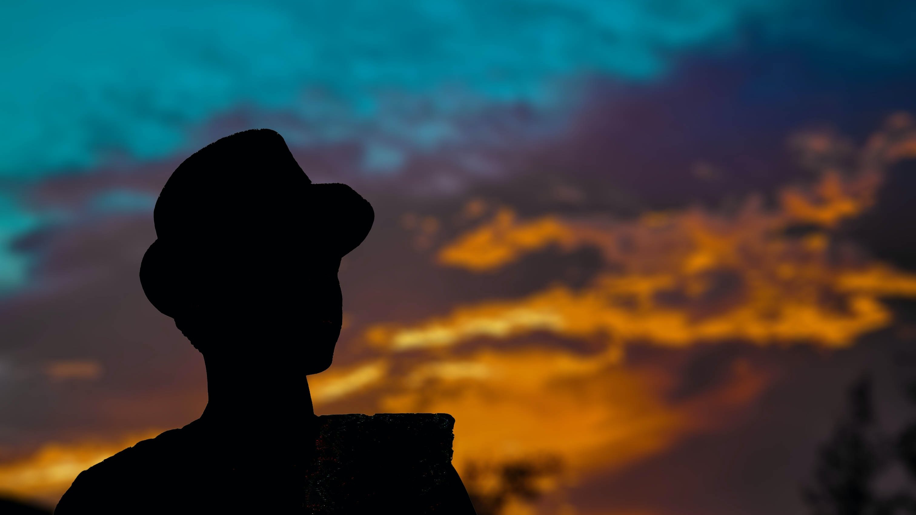 L'image présente une silhouette d'une personne se tenant debout, sur fond d'un ciel coloré au crépuscule. Le personnage porte un chapeau et semble contempler l'horizon. Le ciel est un mélange vibrant de bleu, d'orange et de violet, les nuages créant une atmosphère paisible et dramatique. Les couleurs chaudes du coucher de soleil se marient harmonieusement avec les teintes fraîches du ciel, engendrant une ambiance tranquille et contemplative.