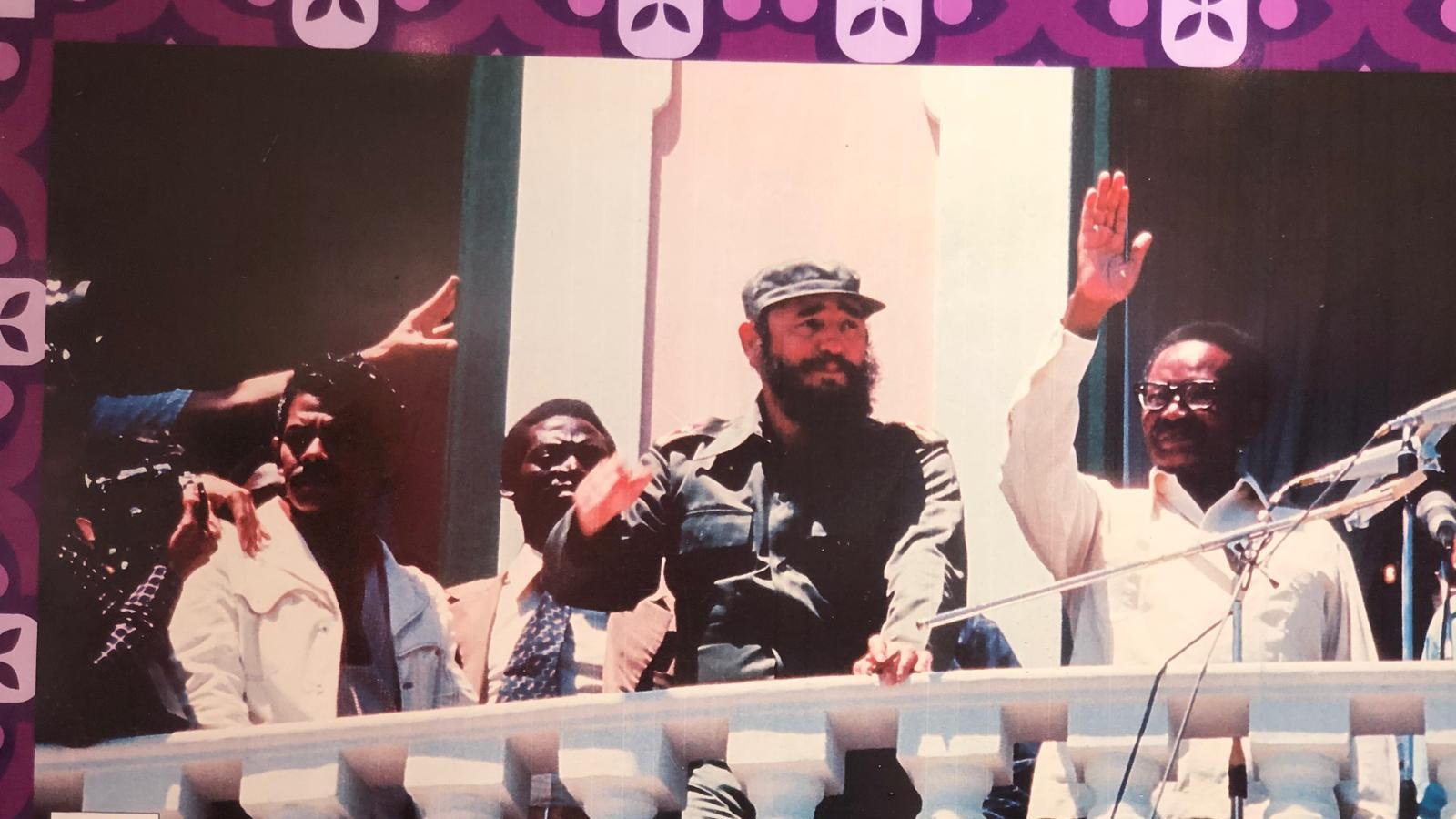 L'image représente un groupe de personnes sur un balcon, probablement lors d'un événement politique. Au centre, un homme avec une barbe, portant un uniforme militaire et un chapeau, lève la main en signe de salutation. À sa gauche, un autre homme en chemise claire semble aussi s'adresser à la foule. À droite, un homme en costume fait un geste similaire. En arrière-plan, on peut voir des murs peints dans des teintes claires, et le décor est encadré par des motifs colorés à la base de l'image. L'atmosphère semble festive et engageante.