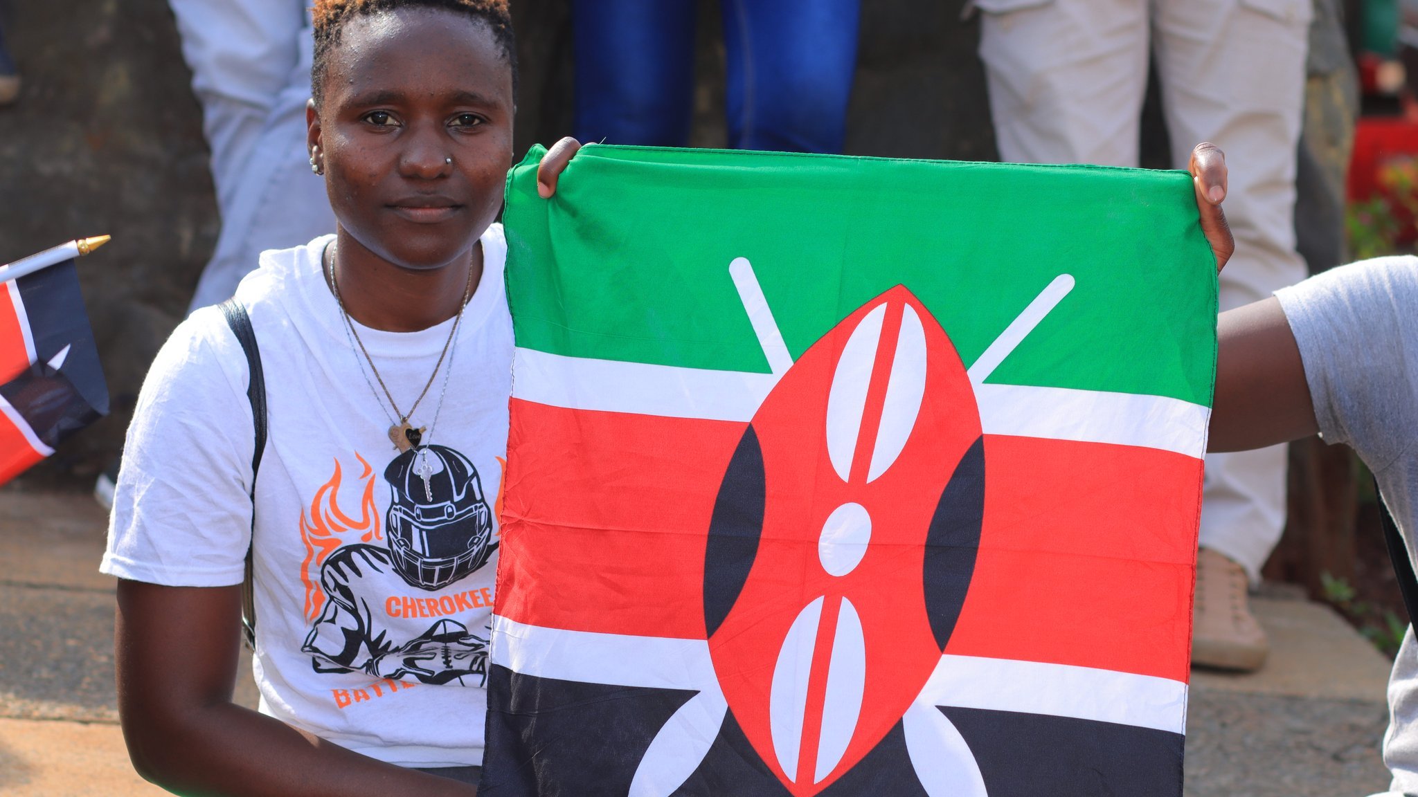 Dans cette image, une jeune femme sourit tout en tenant une grande drapeau du Kenya. Le drapeau est composé de bandes horizontales de couleur noire, rouge et verte, avec un motif central représentant un écu et des flèches. La femme porte un t-shirt blanc orné d'un motif sportif, avec un choker en pendentif autour du cou. En arrière-plan, on peut apercevoir d'autres personnes, certaines tenant également des drapeaux, créant une ambiance festive et patriotique.