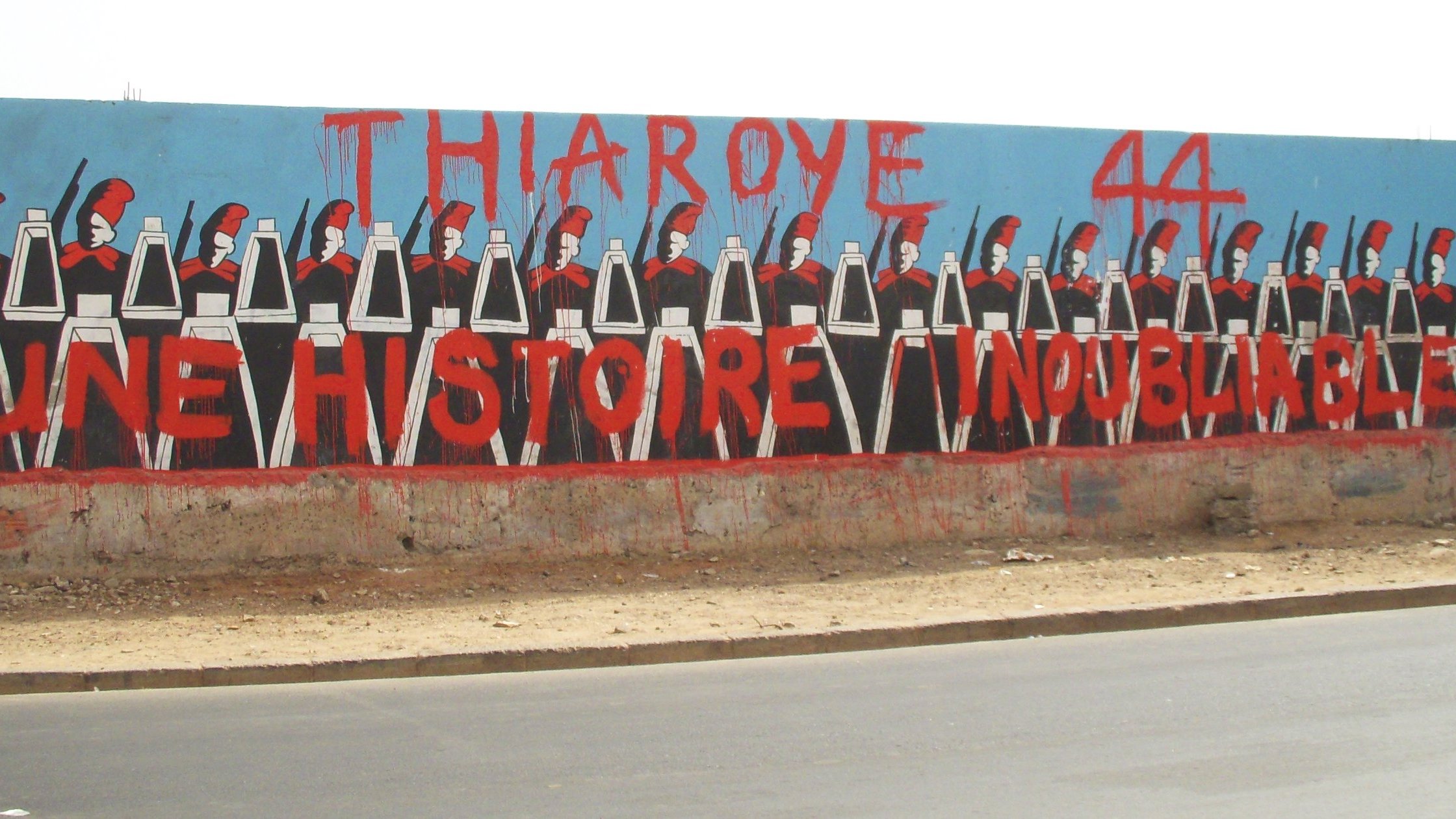 L'image montre une grande fresque murale peinte sur un mur. Au premier plan, on voit une rangée de silhouettes représentant des soldats, chacun portant un uniforme noir et un casque. Ils sont alignés de manière uniforme, les bras le long du corps. En arrière-plan, le mur est peint en bleu, ce qui fait ressortir les personnages. En haut, on peut lire en grandes lettres rouges le texte "THIAROYE 44", et en dessous, "UNE HISTOIRE INOUBLIABLE". Cette œuvre évoque une mémoire historique importante, probablement liée à des événements marquants qui ont eu lieu à Thiaroye, une ville du Sénégal. L'ensemble de l'image crée une ambiance à la fois sérieuse et respectueuse, honorant un passé collectif.