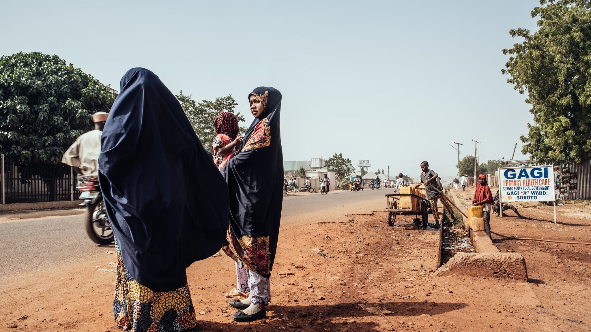 Dans cette image, nous sommes sur une route dans un environnement urbain. Sur le côté gauche, deux femmes portant des vêtements traditionnels de couleur sombre se tiennent ensemble. L'une d'elles a un sourire et semble discuter. Elles sont entourées de terre brune, et l'atmosphère est calme. Sur le côté droit, un homme est assis près d'un petit chariot, tandis qu'un panneau indique « GAGI », signalant un commerce local. On peut apercevoir d'autres personnes se déplaçant à pied ou à bicyclette sur la route, soulignant la vie active de ce lieu. Au fond, des arbres verdoyants ajoutent une touche de nature à l'environnement urbain.