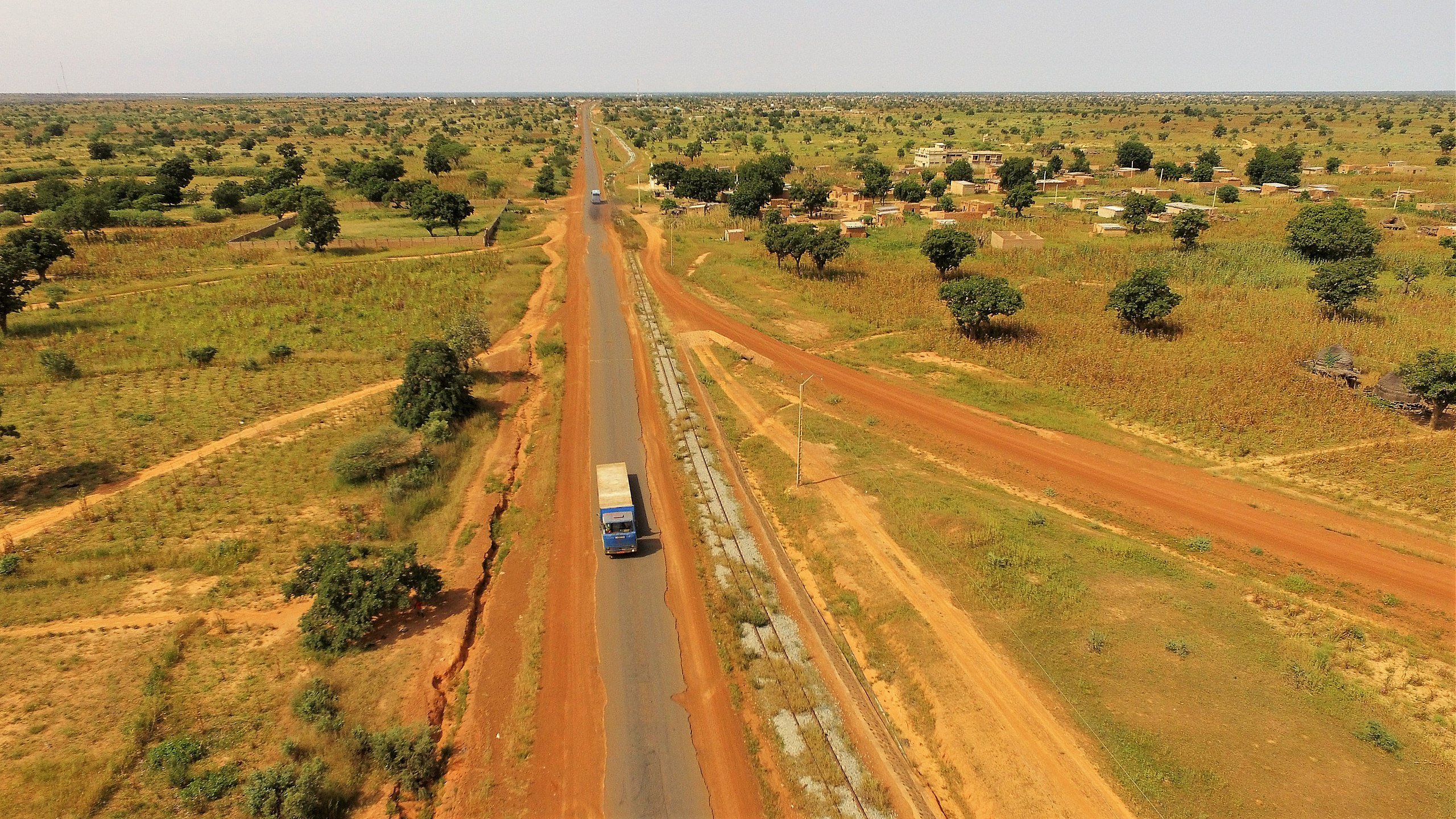 Cette image présente un paysage rural étendu, où une route goudronnée traverse une vaste plaine. Sur la droite, on aperçoit un camion bleu qui circule, indiquant une activité de transport. De part et d'autre de la route, le sol est d'un rouge terreux, typique de certaines régions africaines, semé de petites collines et de végétation éparse, notamment des arbres et des buissons. Sur la gauche de l'image, on peut distinguer des habitations simples, suggérant la présence d'un village. L'arrière-plan est composé de champs et d'arbres, renforçant l'impression de tranquillité et d'espace ouvert. L'ensemble évoque une atmosphère de ruralité, avec une nature généreuse, baignée par une lumière intense.