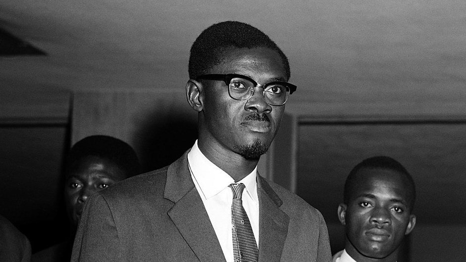 L'image montre un groupe d'hommes en costume, pris dans un cadre formel. Au premier plan, un jeune homme porte des lunettes à monture noire et affiche une expression sérieuse. Il est vêtu d'un costume sombre avec une chemise claire et une cravate. Derrière lui, deux autres hommes sont présents, également en costume, portant des expressions neutres. La lumière est douce, et l'atmosphère dégage un sentiment de gravité ou d'importance. Les murs autour d'eux semblent être peu décorés, mettant l'accent sur les personnes au centre de l'image.