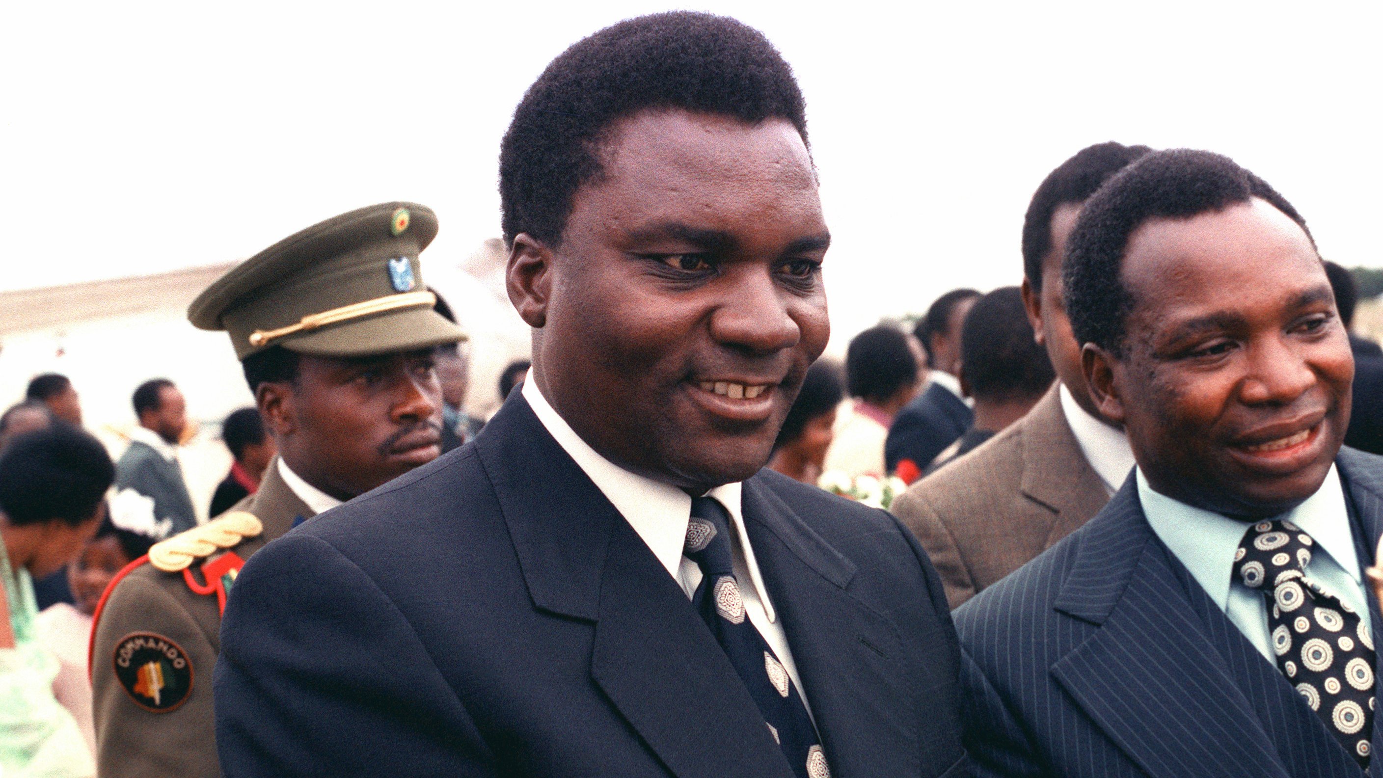 L'image montre un groupe de personnes en extérieur lors d'un événement formel. Au premier plan, deux hommes bien habillés sourient et semblent discuter. L'un porte un costume noir avec une cravate à motifs, tandis que l'autre arbore un costume rayé avec une cravate aux motifs plus délicats. En arrière-plan, un homme en uniforme militaire observe la scène, ce qui ajoute une dimension de solennité à l'événement. Le fond est lumineux, et on peut deviner une atmosphère de convivialité et d'échange entre les participants.
