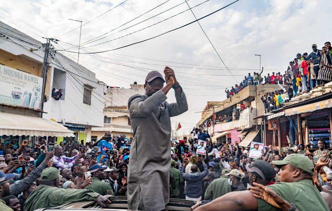 L'image montre une scène dynamique et animée dans une rue bondée. Au centre, un homme, vêtu d'une tenue grise, se tient debout sur un véhicule, levant les bras avec enthousiasme. Les gens autour de lui, une foule importante, applaudissent et expriment leur soutien. On peut entendre des cris de joie et des chants de la part des personnes qui entourent le véhicule. À l'arrière-plan, des bâtiments et des personnes sur les balcons ajoutent à l'atmosphère de ferveur et de célébration. Le ciel est partiellement nuageux, créant un éclairage intéressant sur cette scène vibrante.