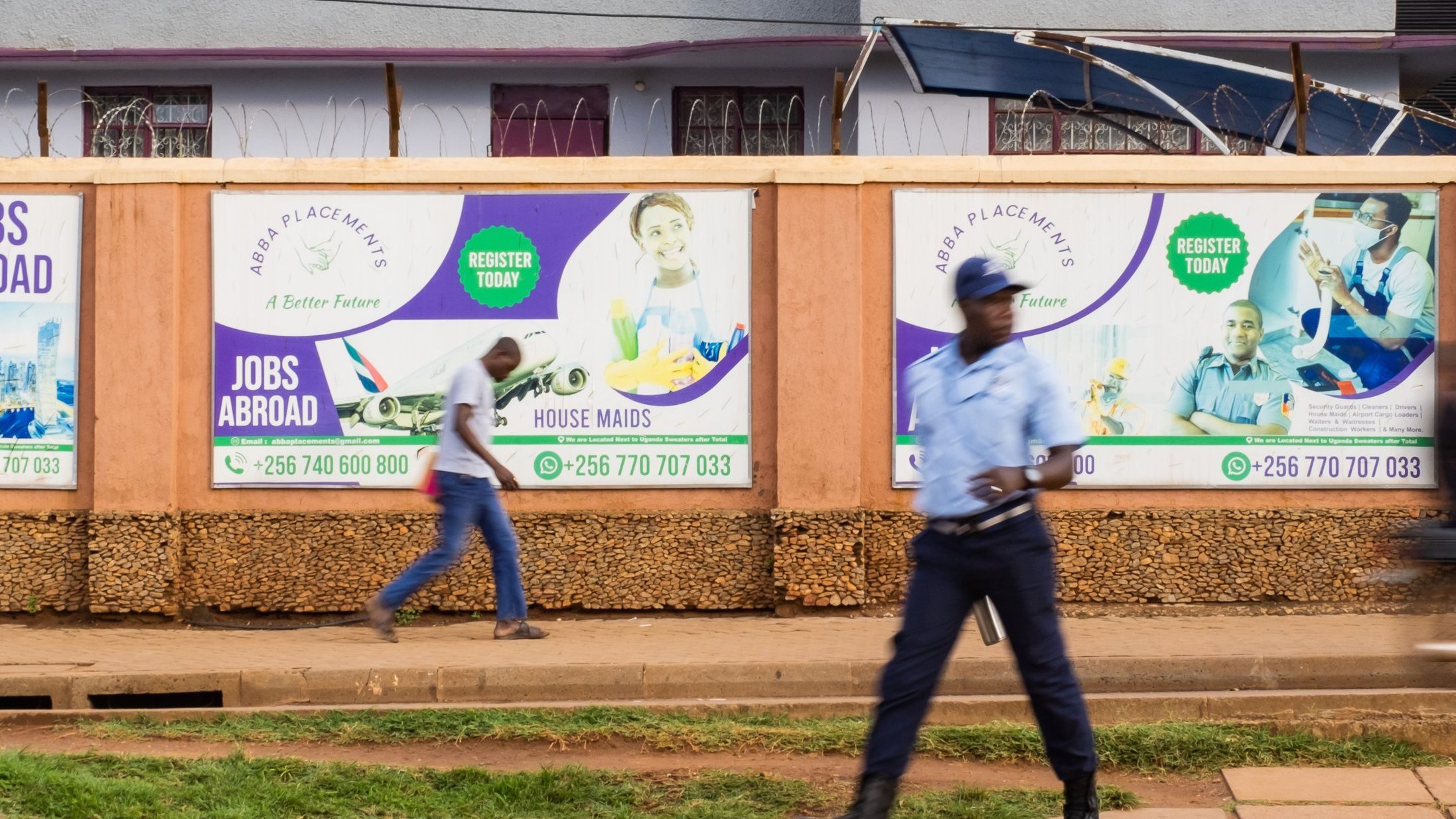 L'image montre un environnement urbain animé. Sur un mur en bois, plusieurs affiches colorées sont accrochées, promouvant des services tels que des opportunités de travail à l'étranger et des services de ménage. Les affiches présentent des visages souriants de personnes, évoquant une atmosphère positive et encourageante. Au premier plan, un homme en uniforme, probablement un agent de sécurité, marche avec assurance sur le trottoir, tandis qu'un autre passant se déplace rapidement derrière lui. Le sol est pavé, et on peut deviner une verdure légère avec une pelouse bien entretenue. L'ensemble évoque une scène de la vie quotidienne dans une ville dynamique.
