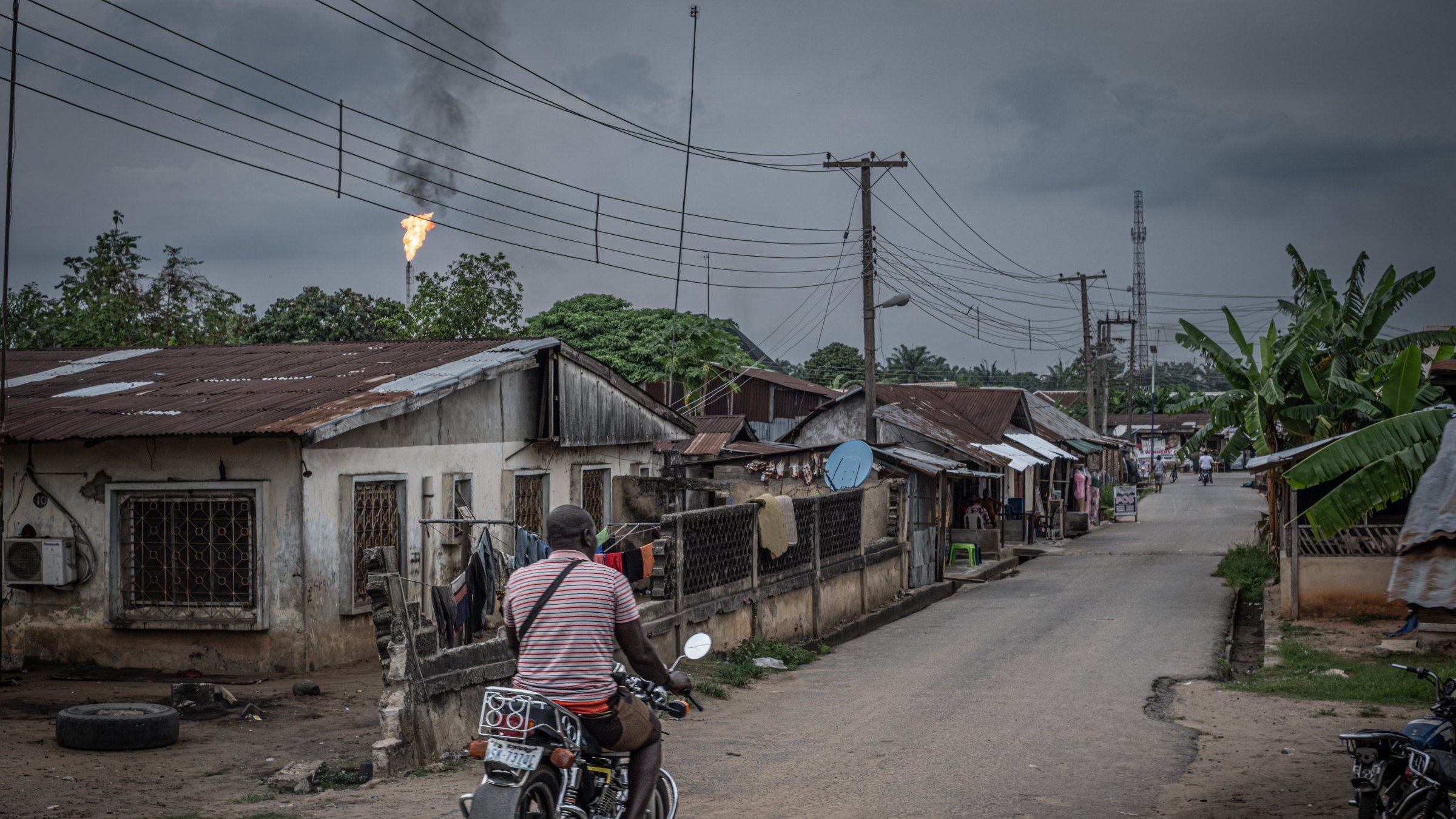 L'image présente une scène urbaine dans un quartier qui semble rural. Une route centrale est bordée de maisons modestes, souvent en tôle, avec des fenêtres grillagées. À gauche, on apperçoit un homme sur une moto, portant un t-shirt rayé, qui se dirige vers l'avant, ajoutant une touche de mouvement à la scène. En arrière-plan, des poteaux électriques sont visibles, ainsi qu'un tuyau de gaz qui rejette une flamme, indiquant peut-être une installation industrielle à proximité. Le ciel est couvert, avec des nuages sombres, créant une atmosphère légèrement mélancolique. À droite, des plantations de bananiers ajoutent une touche de verdure. L'ensemble dépeint une réalité de vie quotidienne dans un environnement à la fois simple et intéressant.
