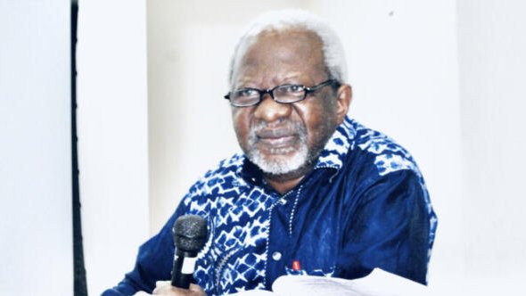 L'image présente un homme assis à une table, tenant un microphone. Il porte des lunettes et a des cheveux gris et bouclés. Son visage exprime une certaine sagesse et réflexion. Il est habillé d'une chemise à motifs, principalement bleue et blanche. Le fond de l'image est neutre, mettant en avant sa présence. On peut sentir une atmosphère de concentration, probablement lors d'un discours ou d'une discussion.