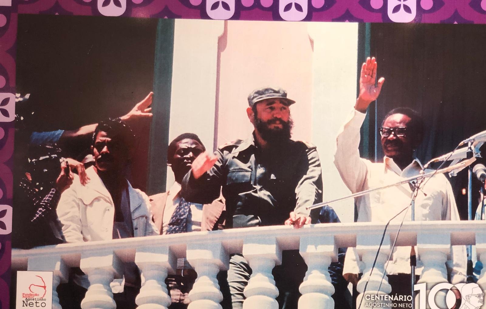L'image représente un groupe de personnes sur un balcon, probablement lors d'un événement politique. Au centre, un homme avec une barbe, portant un uniforme militaire et un chapeau, lève la main en signe de salutation. À sa gauche, un autre homme en chemise claire semble aussi s'adresser à la foule. À droite, un homme en costume fait un geste similaire. En arrière-plan, on peut voir des murs peints dans des teintes claires, et le décor est encadré par des motifs colorés à la base de l'image. L'atmosphère semble festive et engageante.