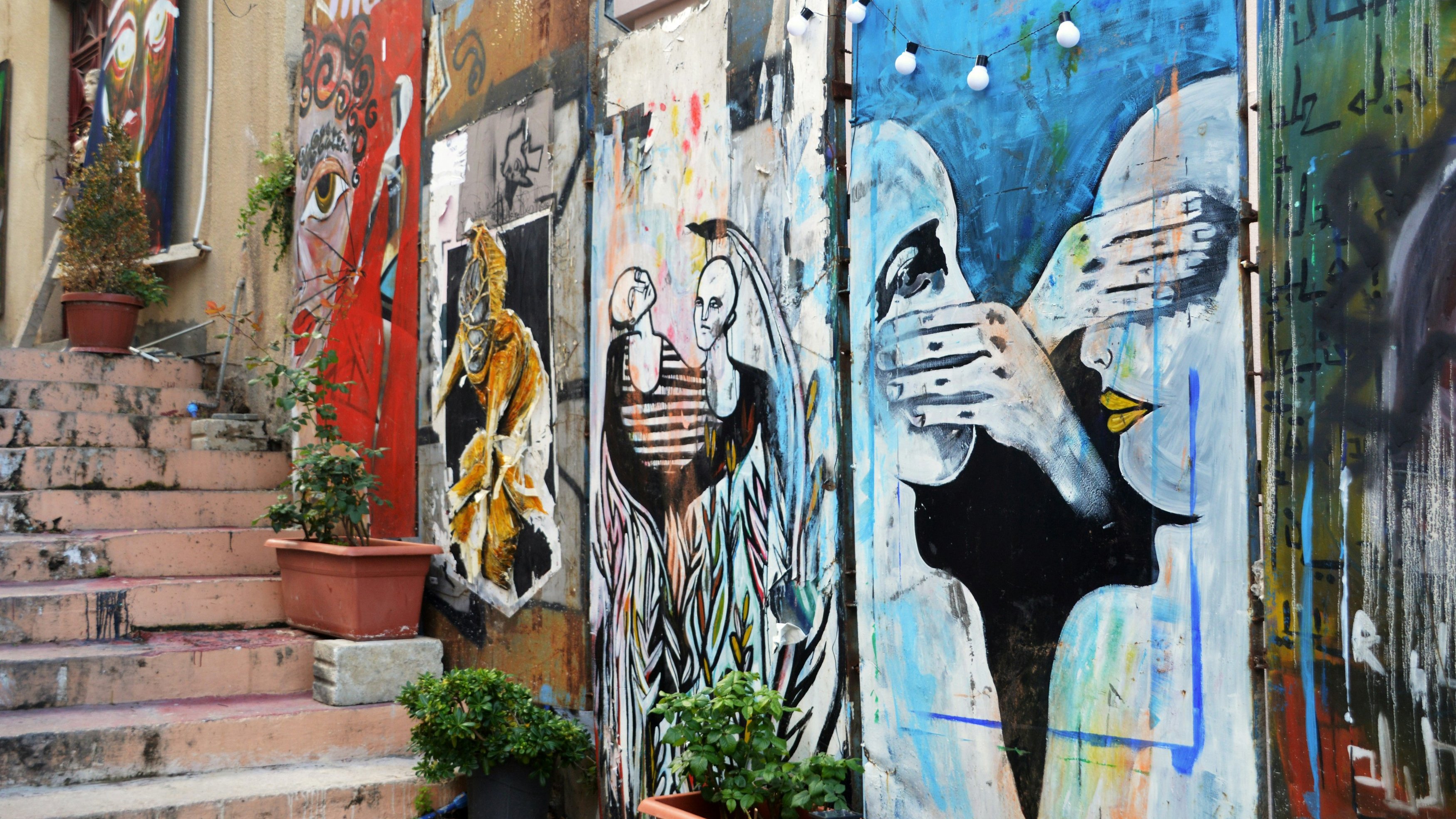 L'image présente un mur de graffitis colorés qui ornent une ruelle. On peut distinguer plusieurs œuvres d'art, chacune ayant son propre style. À gauche, une figure regardante avec des détails réalistes, suivie d'illustrations plus abstraites et expressives au centre et à droite, représentant des visages et des mains en mouvement. Les couleurs dominantes incluent des nuances vives de bleu, d'orange et de noir. Des pots de fleurs, avec des plantes vertes, ajoutent une touche de nature à l'environnement urbain. Au-dessus des œuvres, des guirlandes lumineuses créent une ambiance chaleureuse. Les escaliers en pierre accompagnent le mur, suggérant une connexion avec l'espace environnant. L'ensemble dégage une atmosphère artistique et dynamique, reflet d'un lieu vivant et créatif.