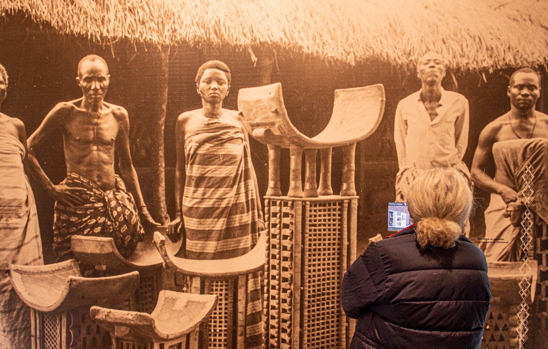 Dans cette image, on aperçoit une scène historique en noir et blanc montrant plusieurs personnes debout, semblant faire partie d'une communauté traditionnelle. Il y a des hommes et des femmes, vêtus de tissus authentiques, avec des poses qui expriment la dignité et la culture. Au centre, on voit des objets artisanaux, comme des chaises ou des tabourets, fabriqués en matériaux naturels, qui portent des motifs décoratifs. À l'arrière-plan, on devine une architecture simple, avec un toit de chaume, créant un cadre authentique. Une femme, de dos, observe cette scène, tenant un appareil dans ses mains, captivée par ce témoignage du passé. L'ambiance générale évoque un sentiment de respect pour les traditions et les histoires des personnes représentées.