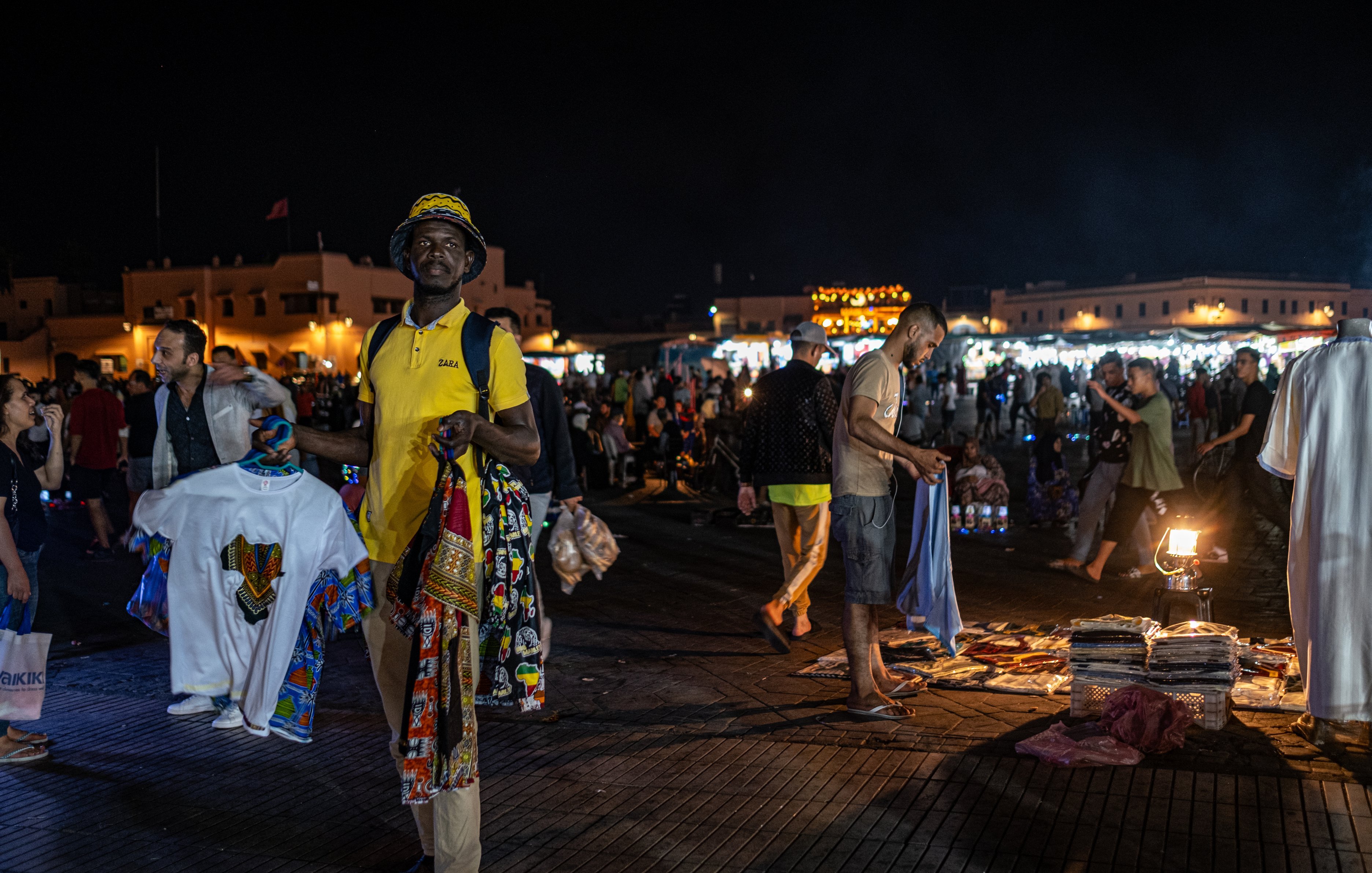 Dans cette image prise la nuit dans un marché animé, on voit un homme au premier plan portant un t-shirt jaune et un chapeau. Il tient plusieurs vêtements, avec des motifs colorés, dans chaque main. L'arrière-plan est rempli de personnes se déplaçant dans le marché, créant une ambiance vivante et dynamique. On peut également apercevoir des lumières brillantes provenant de stands qui éclairent la scène, ajoutant à l'animation nocturne du lieu. La texture du sol semble être en bois ou en pavé, créant un environnement chaleureux. L'air est rempli des sons de conversations et de l'agitation du marché.