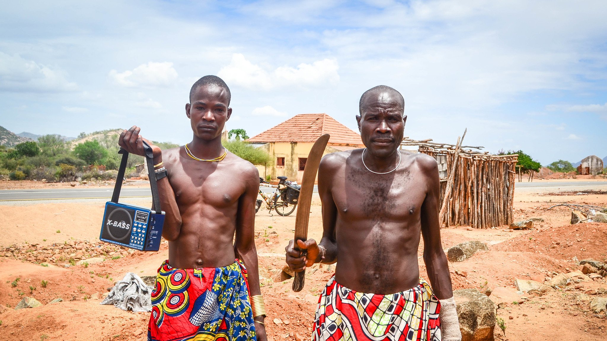 L'image montre deux hommes debout sur un terrain sec et rocheux. Ils portent des vêtements colorés avec des motifs vibrants, typiques d'une culture africaine. Le premier homme, à gauche, tient un poste de radio portable, tandis que le second, à droite, brandit une grande machette. En arrière-plan, on peut apercevoir des bâtiments simples, ainsi qu'une nature aride avec quelques arbres. Le ciel est bleu avec quelques nuages. L'atmosphère semble calme, mais la présence des objets et des vêtements indique une culture dynamique et vivante.
