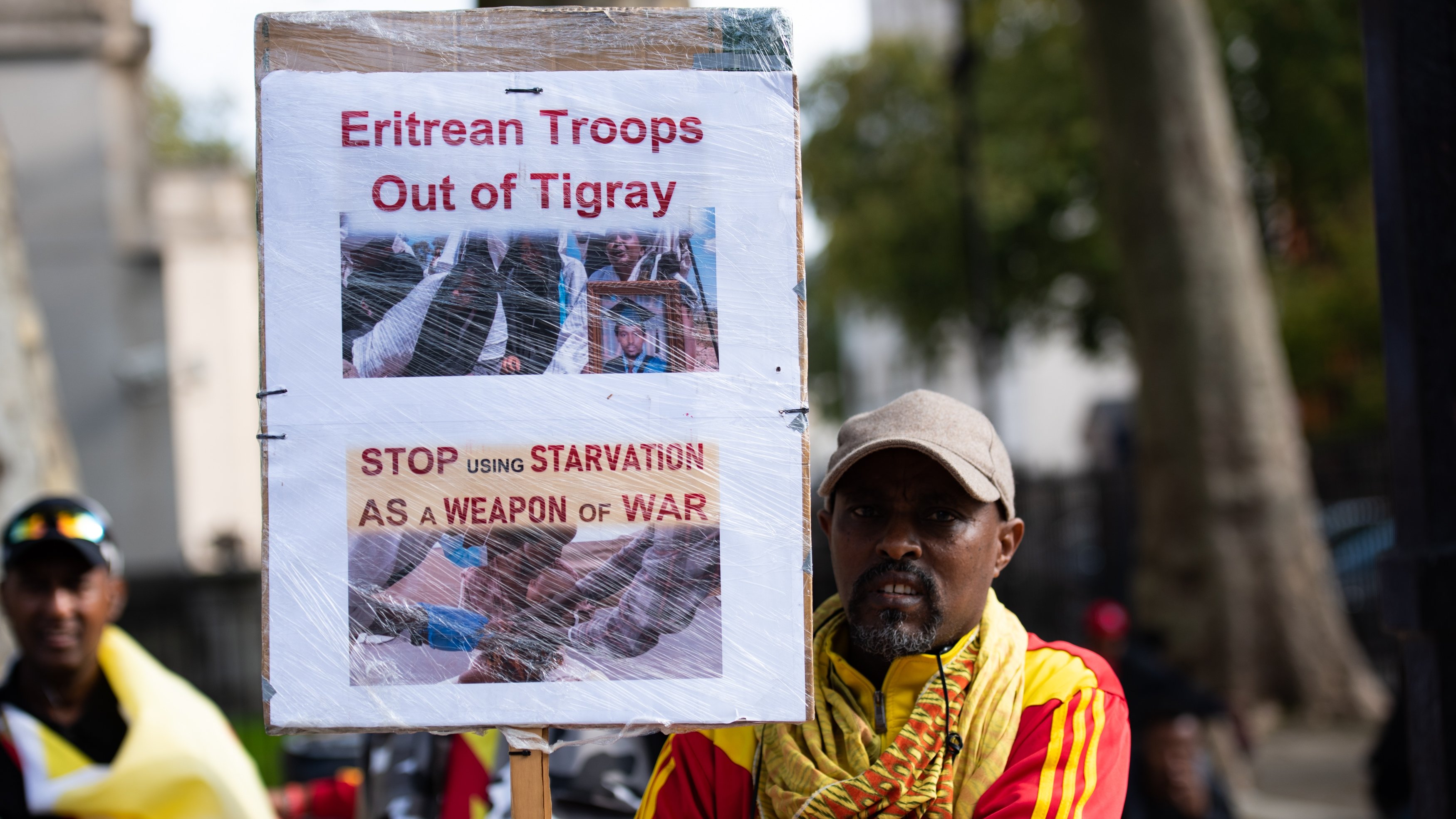 L'image montre un homme qui tient une pancarte haute, sur laquelle sont affichées des images et des textes en anglais. La pancarte exprime des revendications concernant le retrait des troupes érythréennes de la région du Tigré, en Éthiopie. Les mots en gras, "Eritrean Troops Out of Tigray" et "STOP USING STARVATION AS A WEAPON OF WAR", soulignent un appel urgent pour mettre fin à la souffrance causée par la guerre. L'homme porte un chapeau gris et est vêtu d'une écharpe colorée, probablement aux couleurs de l'Éthiopie, ce qui suggère un engagement émotionnel et une participation à une manifestation. En arrière-plan, on peut apercevoir d'autres personnes qui semblent partager des préoccupations similaires. L'ambiance est chargée d'émotion et de détermination.