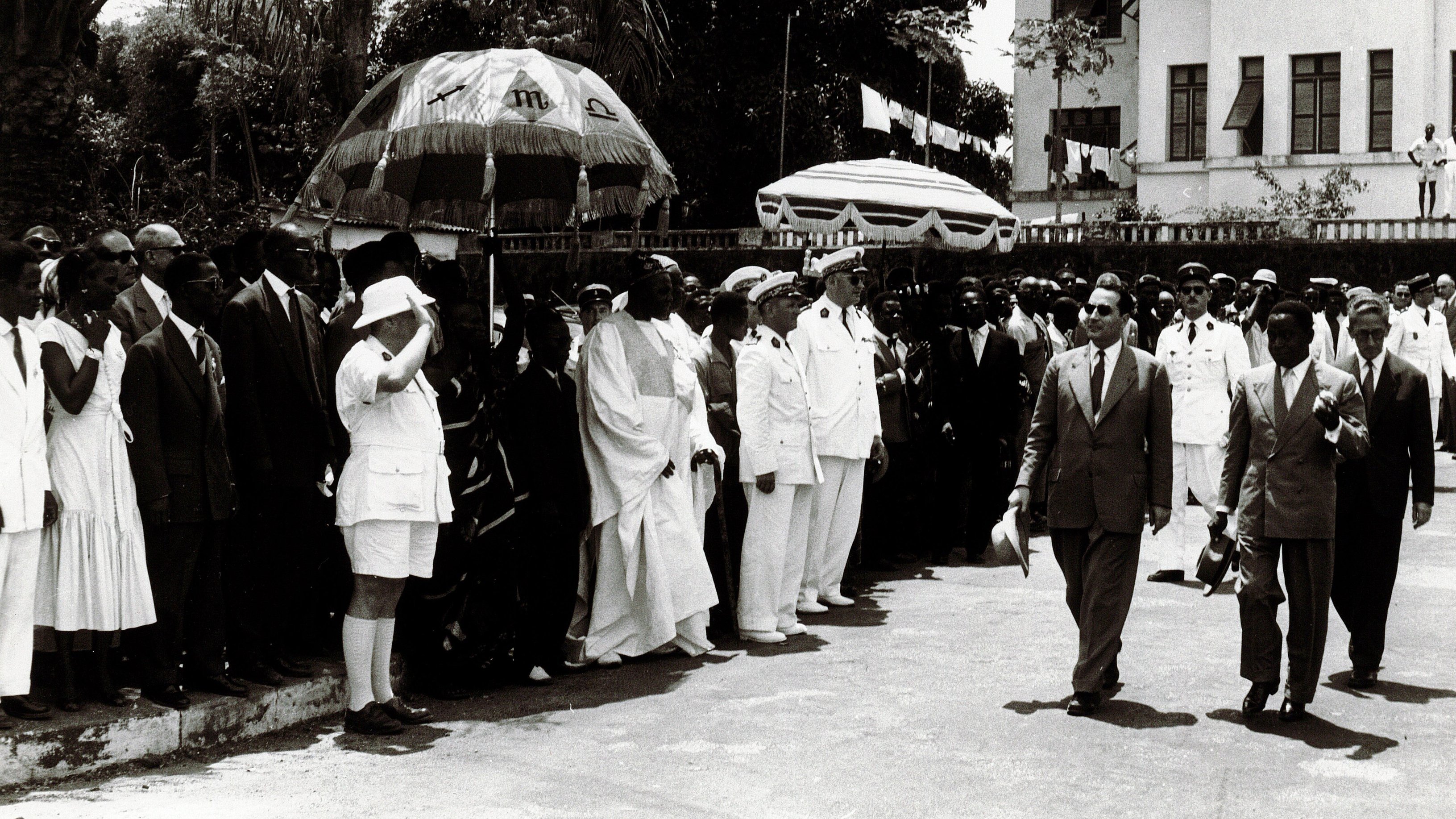 L'image montre un groupe de personnes rassemblées en extérieur, probablement lors d'un événement officiel. Au centre, des hommes en costumes formels marchent, entourés de dignitaires et de membres du public. Certains portent des uniformes militaires, tandis que d'autres sont habillés de manière civile. Il y a également des parapluies décoratifs en arrière-plan. L'atmosphère semble solennelle, et l'environnement suggère une atmosphère tropicale avec des arbres en arrière-plan. La scène donne une impression de cérémonie et de célébration.