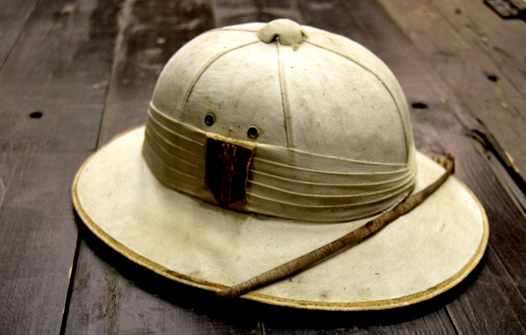 L'image montre un chapeau de style explorer, souvent associé aux expéditions dans des régions chaudes ou tropicales. Le chapeau est de couleur claire, presque beige, avec une large bordure qui protège du soleil. Sa partie supérieure est légèrement surélevée, indiquant une forme arrondie. À l'avant, il y a une bande de tissu plissé avec un morceau de bois ou de cuir, donnant un aspect rustique. Une fine tige est posée sur le chapeau, ajoutant une touche naturelle. La surface du chapeau semble légèrement usée, témoignant d'un usage antérieur. Le fond sur lequel il repose est en bois, ce qui renforce l'atmosphère d'aventure.