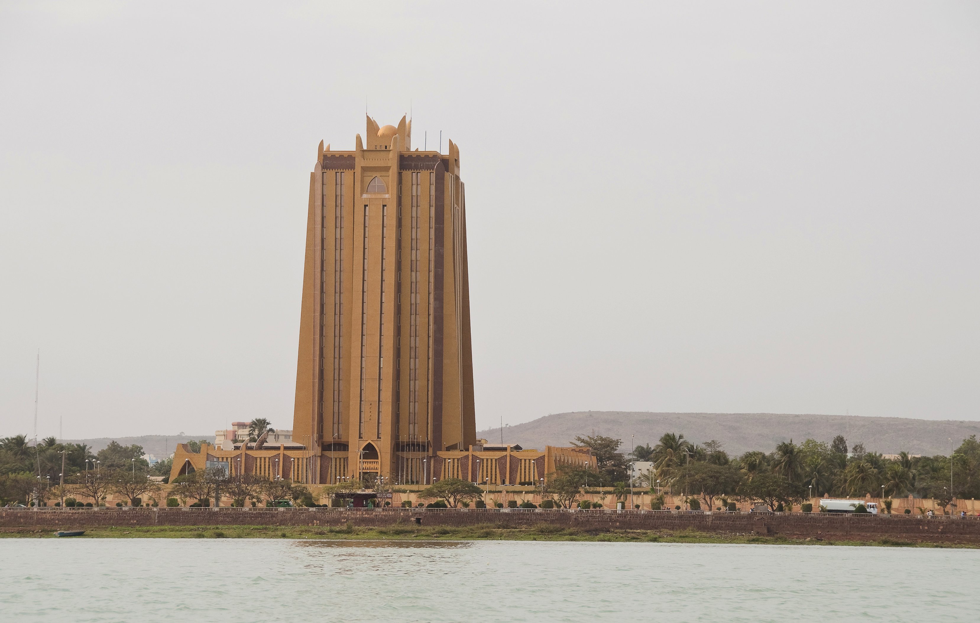 L'image montre un bâtiment imposant qui semble être une tour, située près de l'eau. Ce bâtiment, d'une teinte dorée, a une architecture moderne et élancée. Sa structure se soulève vers le ciel, avec des lignes verticales marquées et des détails décoratifs au sommet, donnant une impression de grandeur. En arrière-plan, on aperçoit quelques collines et des arbres, tandis qu'au premier plan, on voit la surface calme de l'eau. Le ciel est légèrement nuageux, créant une ambiance tranquille et légèrement brumeuse autour de cette scène urbaine.