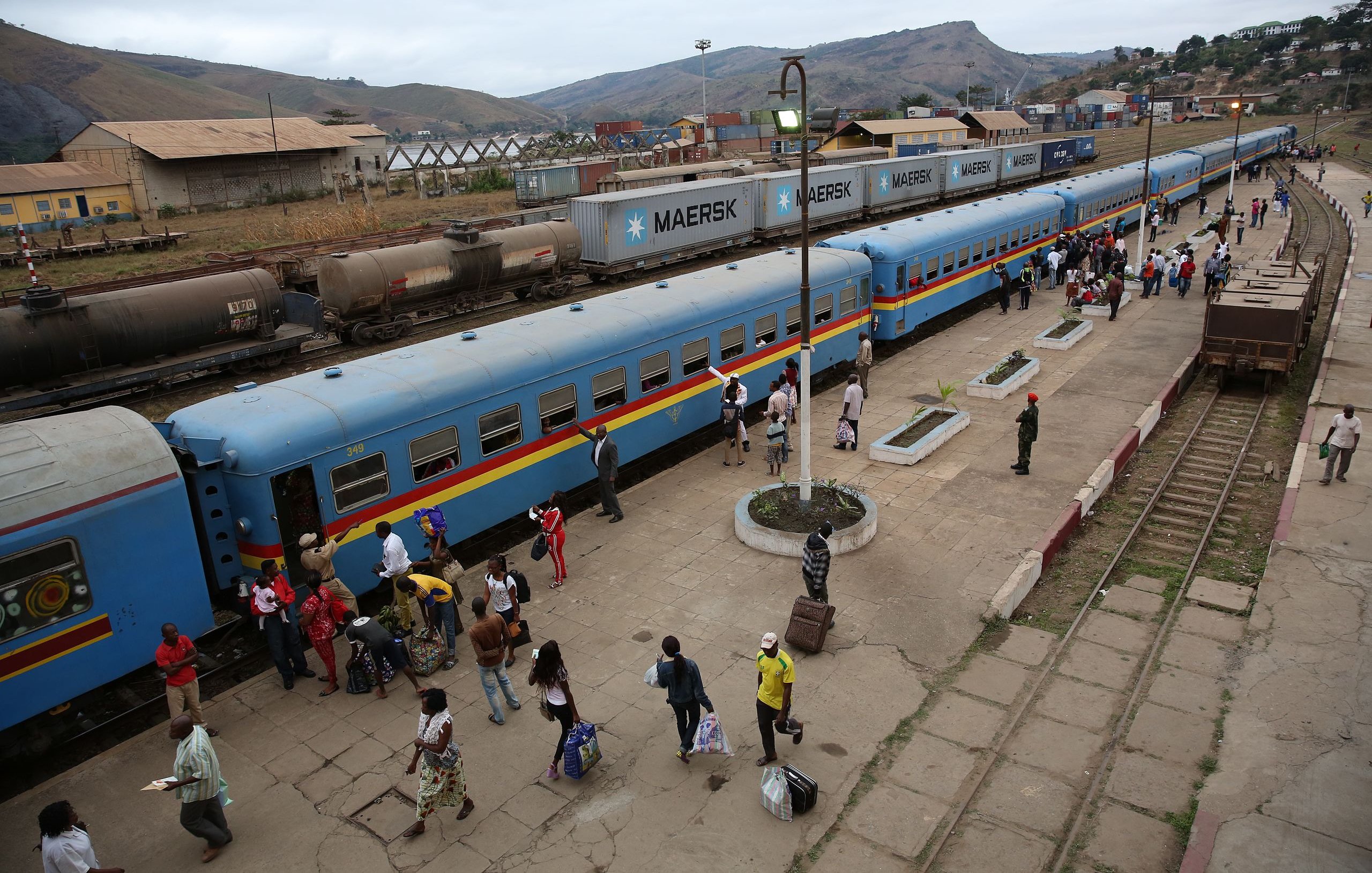 L'image montre une gare animée. De longs trains de marchandises et de passagers sont alignés sur les quais. Les wagons sont d'un bleu vif avec des bandes colorées. Des voyageurs descendent et montent à bord, portant des sacs et des effets personnels. On peut entendre des conversations, des annonces de train, et le bruit des roues sur les rails. En arrière-plan, on aperçoit des collines qui entourent la gare. L'atmosphère est vive et dynamique, avec un mélange de mouvements et d'interactions entre les passagers.