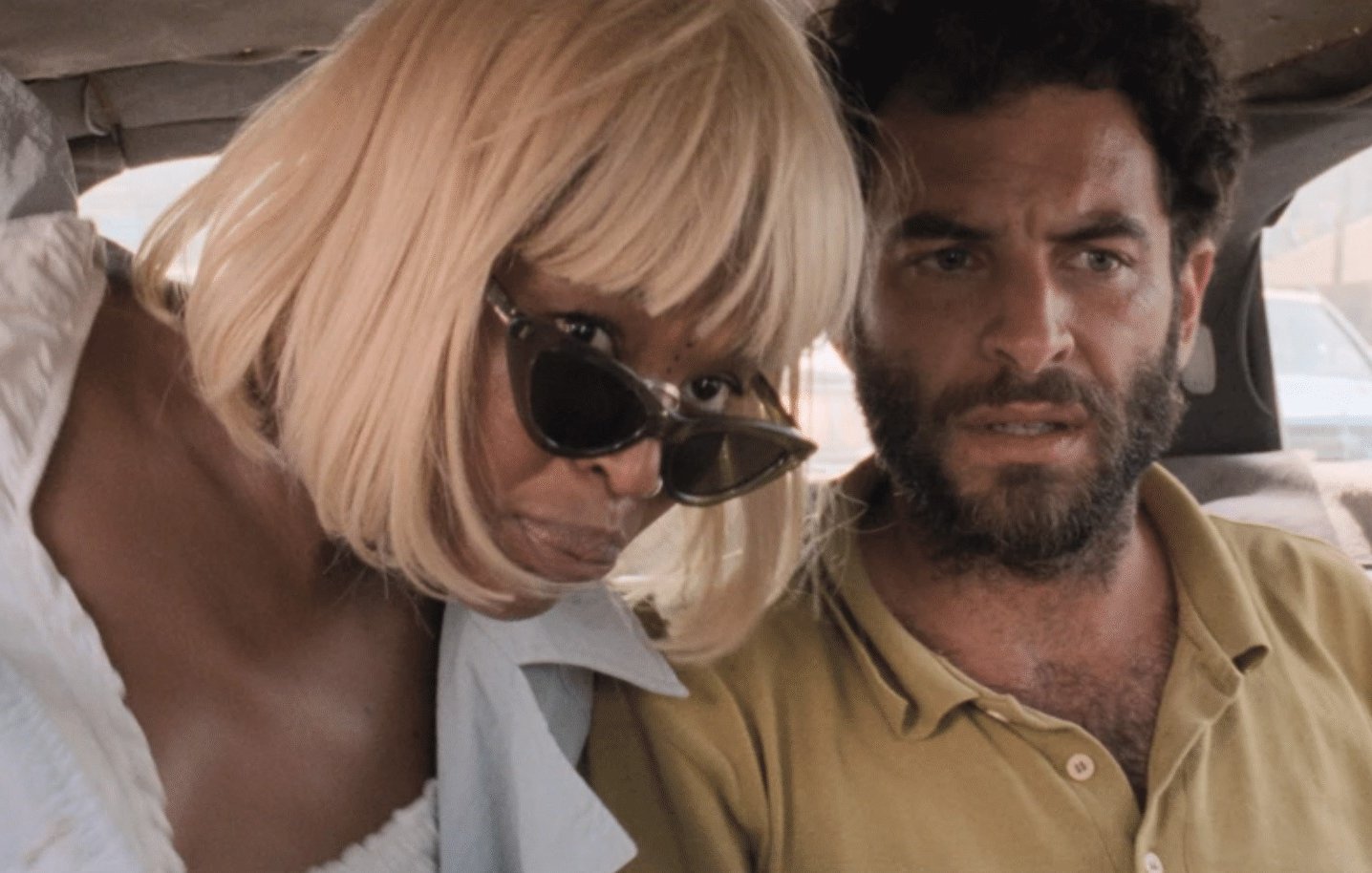 Dans cette image, on voit deux personnes assises à l'arrière d'un véhicule. À gauche, une femme d'une trentaine d'années a les cheveux blonds et courts, coiffés en une coupe élégante. Elle porte de grandes lunettes de soleil noires et une tenue légère. Son expression est attentive, presque inquisitrice. À droite, un homme d'une trentaine d'années également, a une barbe foncée et des cheveux bouclés. Il est vêtu d'une chemise de couleur terre. Son regard est préoccupé et intense, comme s'il était en train de réfléchir à quelque chose d'important. L'atmosphère dans l'image semble suggérer une conversation sérieuse ou un moment de tension partagée. Les sièges du véhicule sont en cuir ou en tissu sombre, et on peut apercevoir des objets non identifiables, peut-être des sacs ou des vêtements, à leurs pieds.