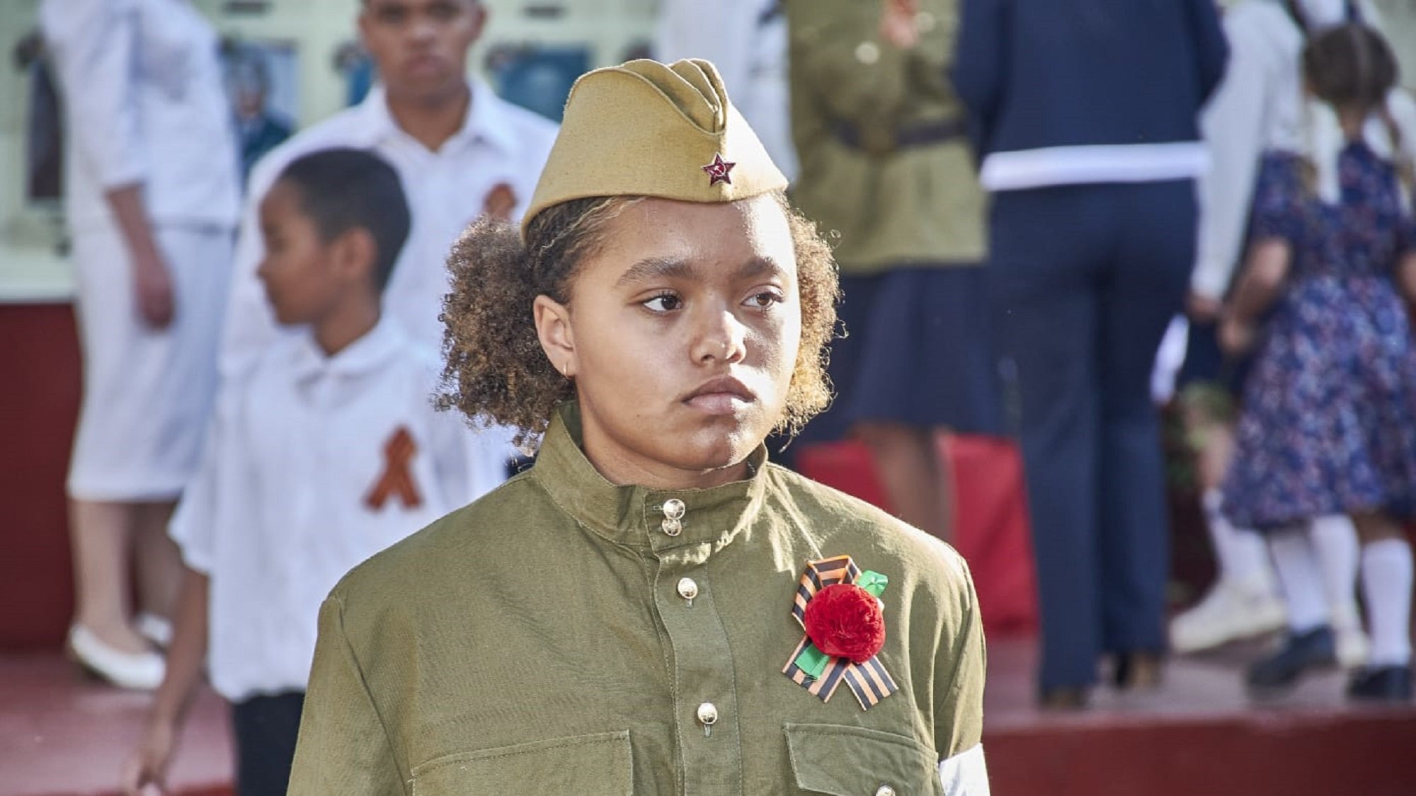 L'image montre une jeune fille portant un uniforme militaire, qui est de couleur olive et a des ornements spécifiques. Elle porte un chapeau similaire, typique des uniformes militaires. Sur sa poitrine, elle affiche une décoration, probablement une médaille ou un insigne, ainsi qu'une fleur rouge. En arrière-plan, on aperçoit d'autres personnes, dont des enfants en vêtements blancs et d'autres habits festifs, suggérant une célébration ou un événement commémoratif. L'expression de la jeune fille semble sérieuse, soulignant l'importance du moment. Le cadre est marqué par une ambiance solennelle, car plusieurs personnes semblent participer à une cérémonie.