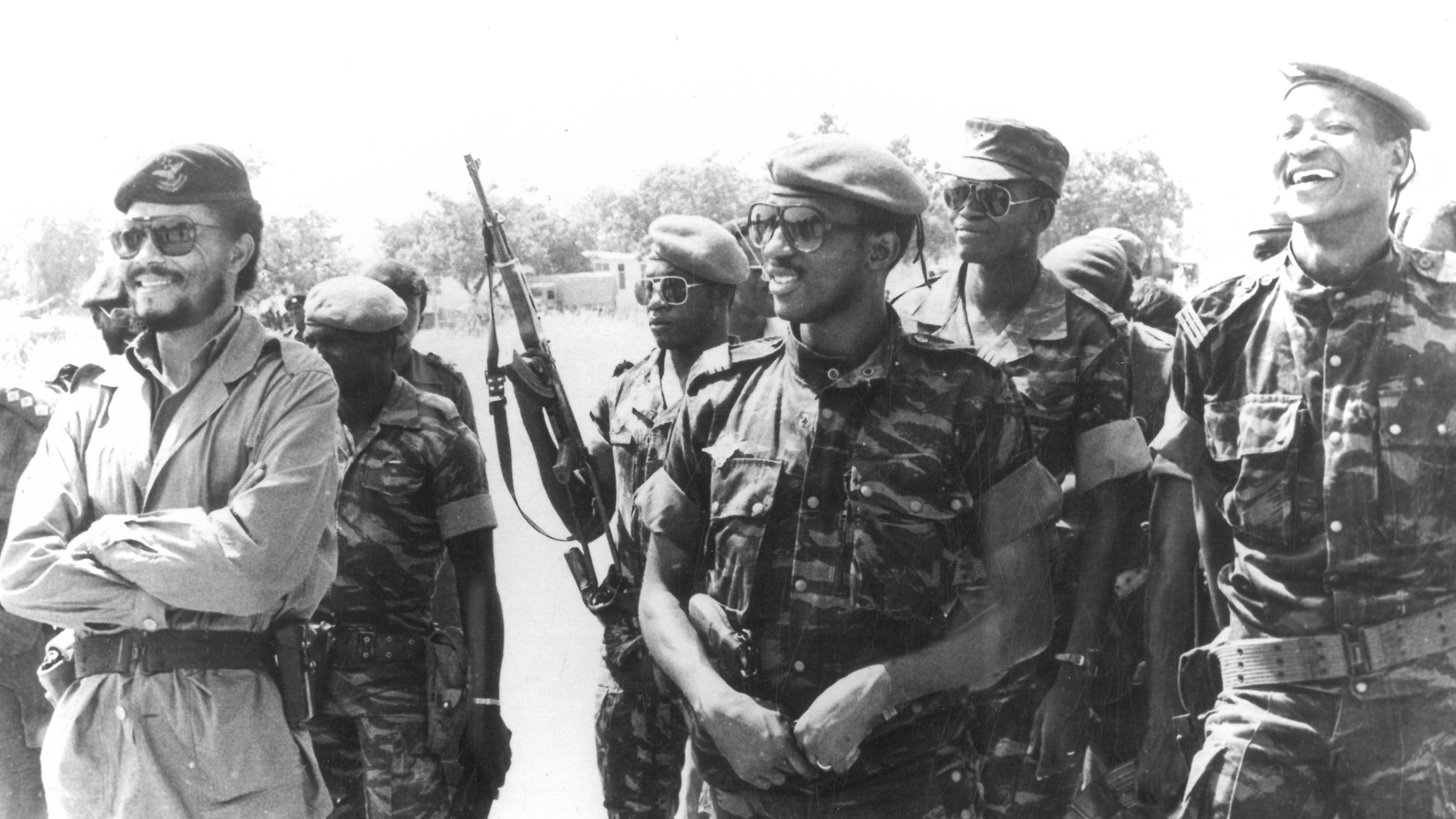 Cette image en noir et blanc montre un groupe de soldats en tenue militaire. Au premier plan, un homme souriant, portant un béret et une tenue de camouflage, se tient à gauche. À sa droite, plusieurs autres soldats, également en uniforme, affichent des expressions de camaraderie. L'un d'eux porte des lunettes de soleil et un autre semble être en train de rire. En arrière-plan, on peut voir des armes accrochées en position debout. L'ambiance générale de l'image suggère une atmosphère de camaraderie et de cohésion parmi les soldats.