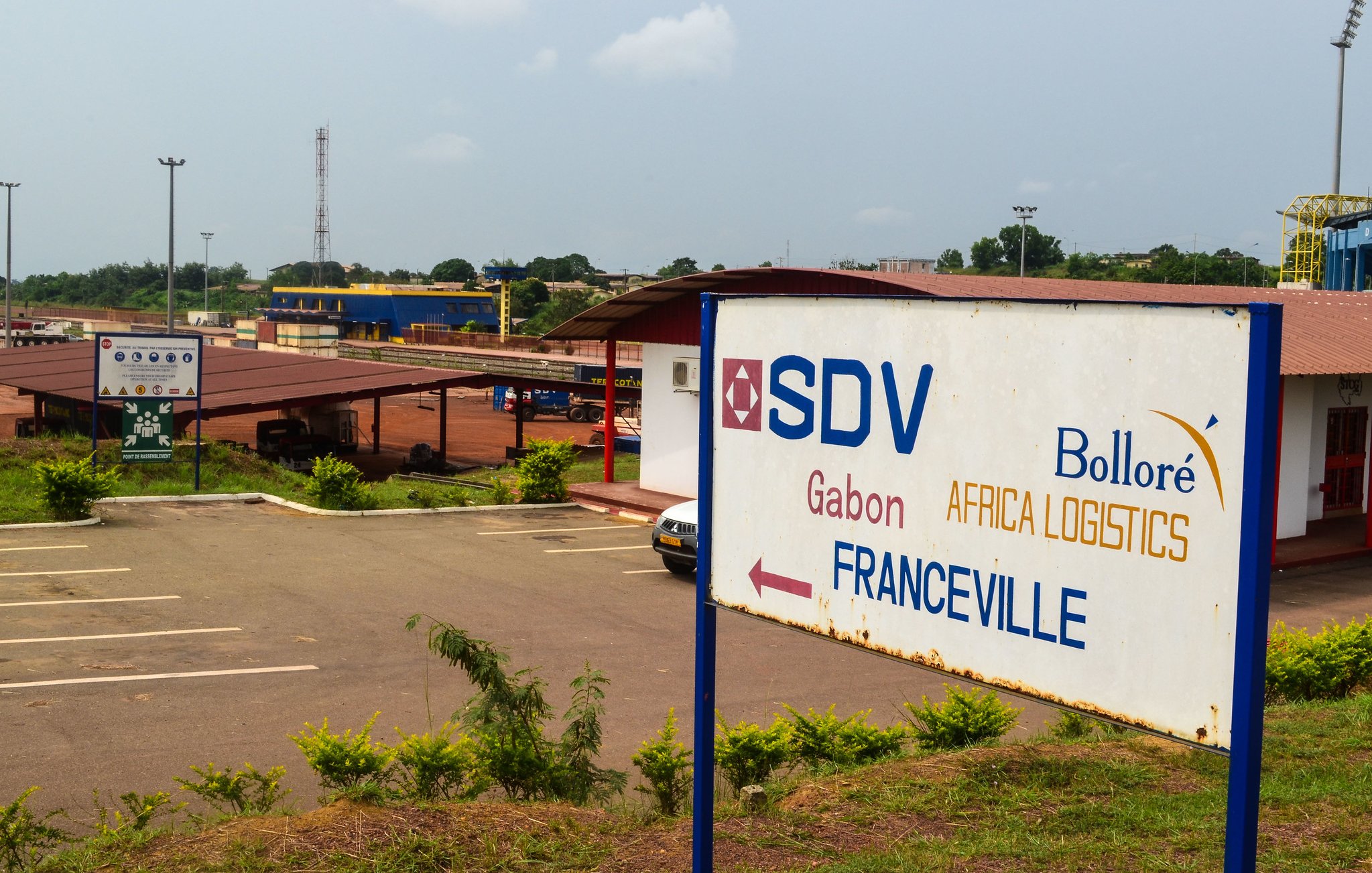 L'image montre un panneau indicatif dans une zone logistique. Ce panneau, situé devant un bâtiment, indique la direction vers Franceville, une ville du Gabon. On peut apercevoir le logo de SDV, ainsi que celui de Bolloré Africa Logistics. Le paysage environnant est assez verdoyant, avec plusieurs arbres et buissons. Au loin, on distingue une infrastructure de transport, possiblement liée à la logistique. L'atmosphère est calme, reflétant un lieu dédié aux activités commerciales et de logistique.