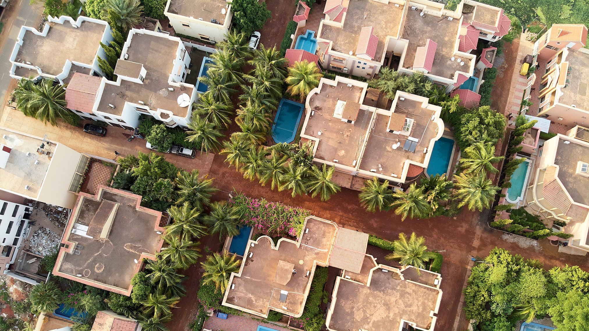L'image montre une vue aérienne d'un complexe résidentiel. On y voit plusieurs maisons aux toits plats, disposées autour de petites piscines bleues. Parmi ces maisons, il y a de nombreuses palmiers qui ajoutent une touche de verdure. Les allées sont en terre et les maisons sont agencées de manière à créer un réseau urbain harmonieux. Les couleurs de la terre et des toits se marient pour donner une ambiance chaleureuse et accueillante au paysage.