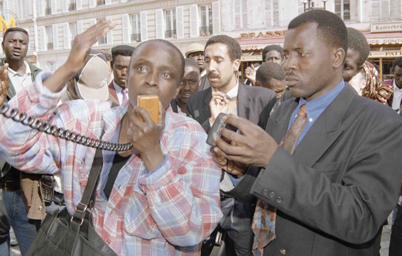 L'image montre un groupe de personnes rassemblées dans un espace public animé. Au premier plan, deux hommes attirent l'attention. L'un d'eux, portant une chemise à carreaux et tenant un microphone, semble s'exprimer avec passion, les bras levés, signalant une forte émotion. L'autre homme, en costume sombre, tient un appareil photo, observant et capturant le moment. En arrière-plan, on peut apercevoir une foule diverse, des visages attentifs, écoutant ou réagissant à la situation. L'atmosphère générale est dynamique, avec un sentiment de mobilisation ou de protestation.