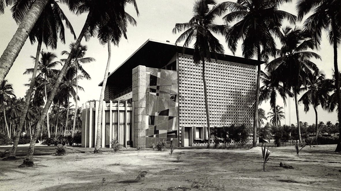 L'image montre un bâtiment moderne, avec une architecture distincte et géométrique. Le mur de la façade est ornée de motifs en relief, créant un jeu d'ombres et de lumière. Des colonnes blanches se dressent devant l'entrée principale, ajoutant une touche de classicisme à la structure. Autour du bâtiment, des palmiers élancés se balancent doucement, donnant une ambiance tropicale. Le sol est nu, avec une végétation assez faible, ce qui permet de mettre en valeur l'architecture. La scène semble baignée dans une lumière naturelle, évoquant une atmosphère calme et sereine.